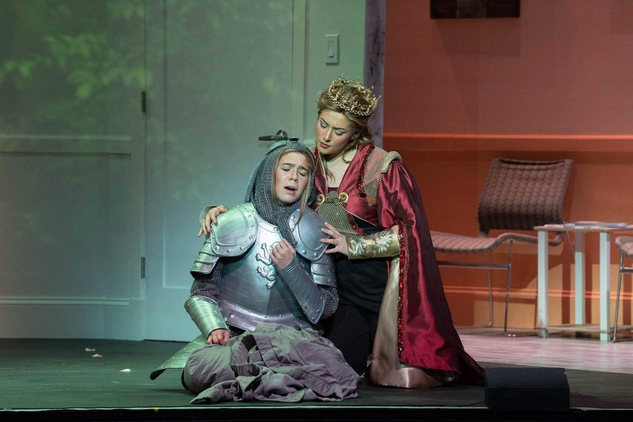 Two women dressed as royalty and a knight with armor on stage during a theatrical performance. The woman in a velvet dress with a crown appears to be comforting the kneeling woman in armor, who is visibly emotional.
