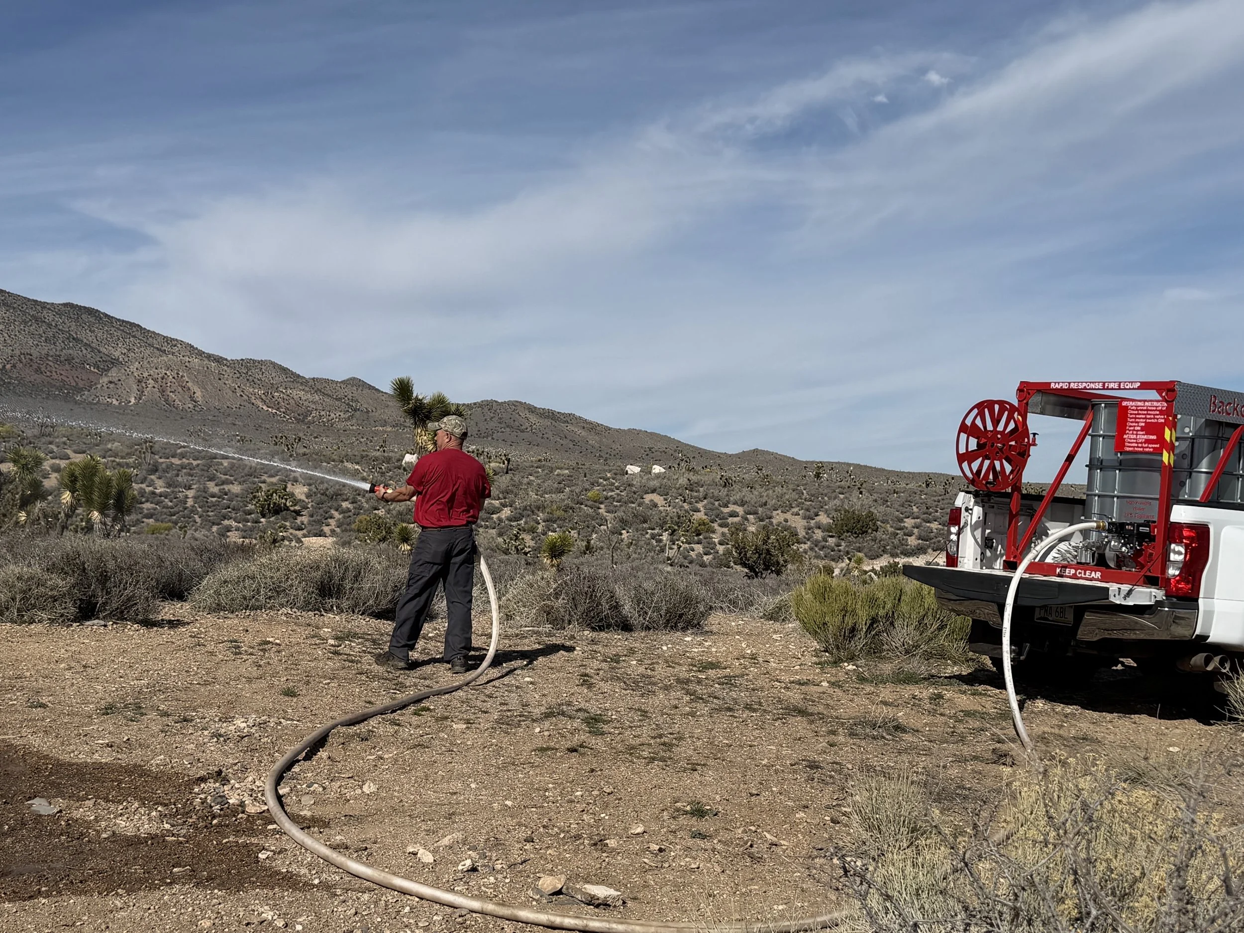 A person in a red shirt and black pants spraying water from a hose on a desert landscape with hills and sparse vegetation. A fire truck with a hose reel is parked nearby.