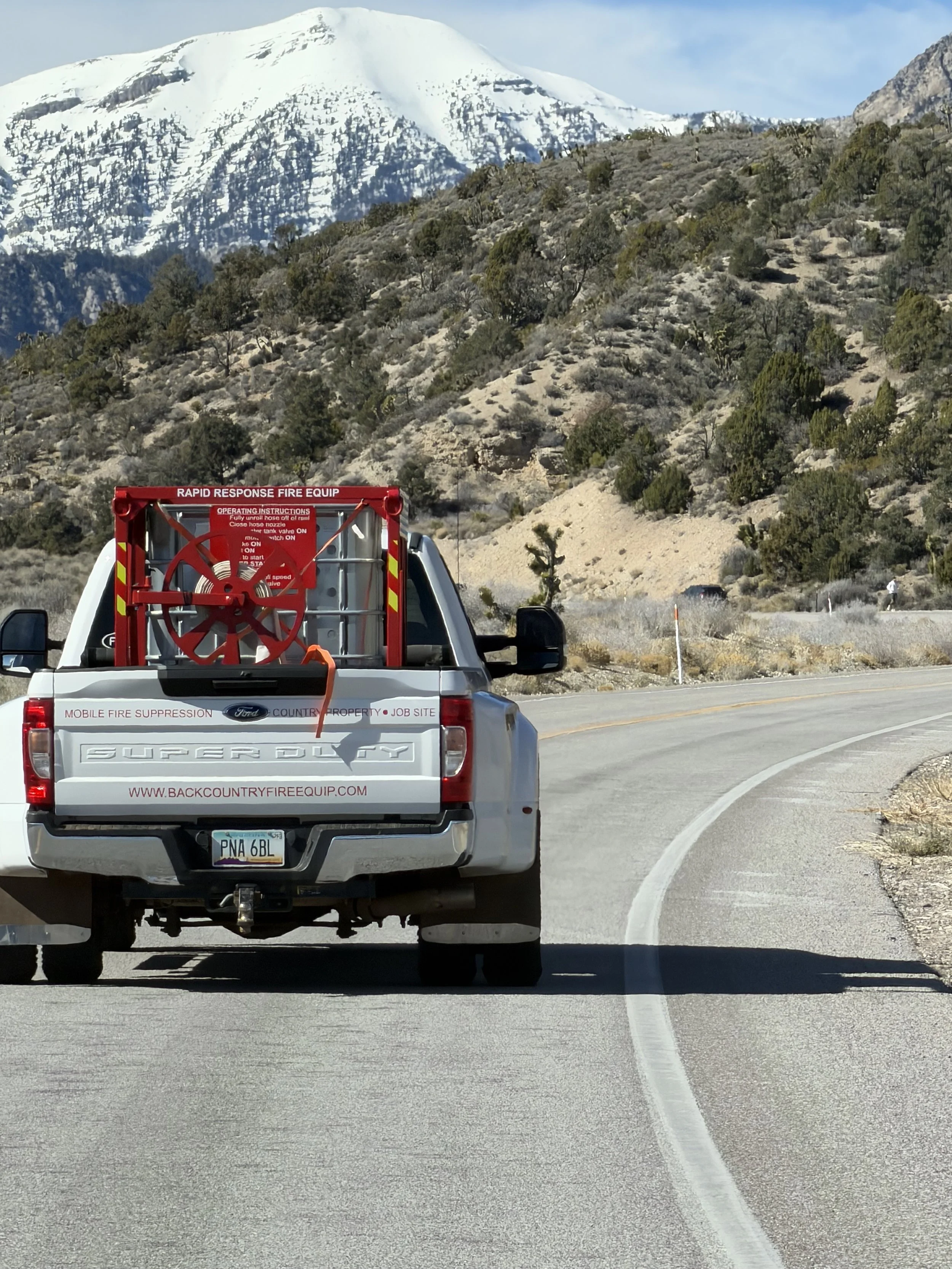 White Ford Super Duty pickup truck with fire suppression equipment in the truck bed, driving on a winding mountain road with snow-capped peaks and rocky hillsides in the background.