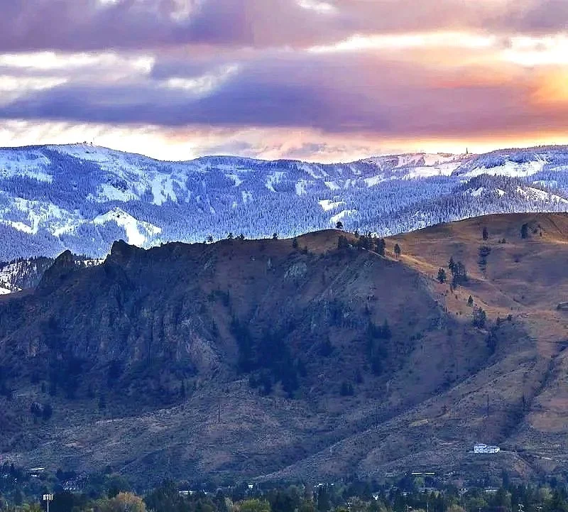 Photo of a mountainous landscape during sunset, with dark rocky hills in the foreground and snow-capped mountains in the background under a colorful cloudy sky.