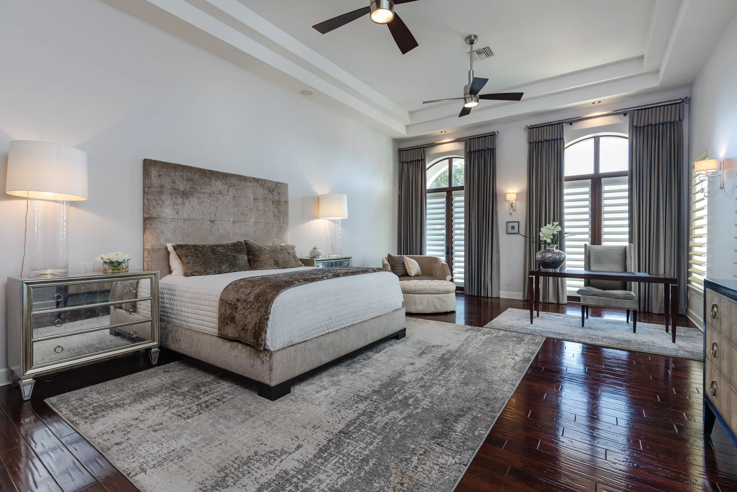 Elegant bedroom with a large bed, mirrored nightstands, armchair, desk with chair, dark wood floors, large windows with curtains, and ceiling fans.