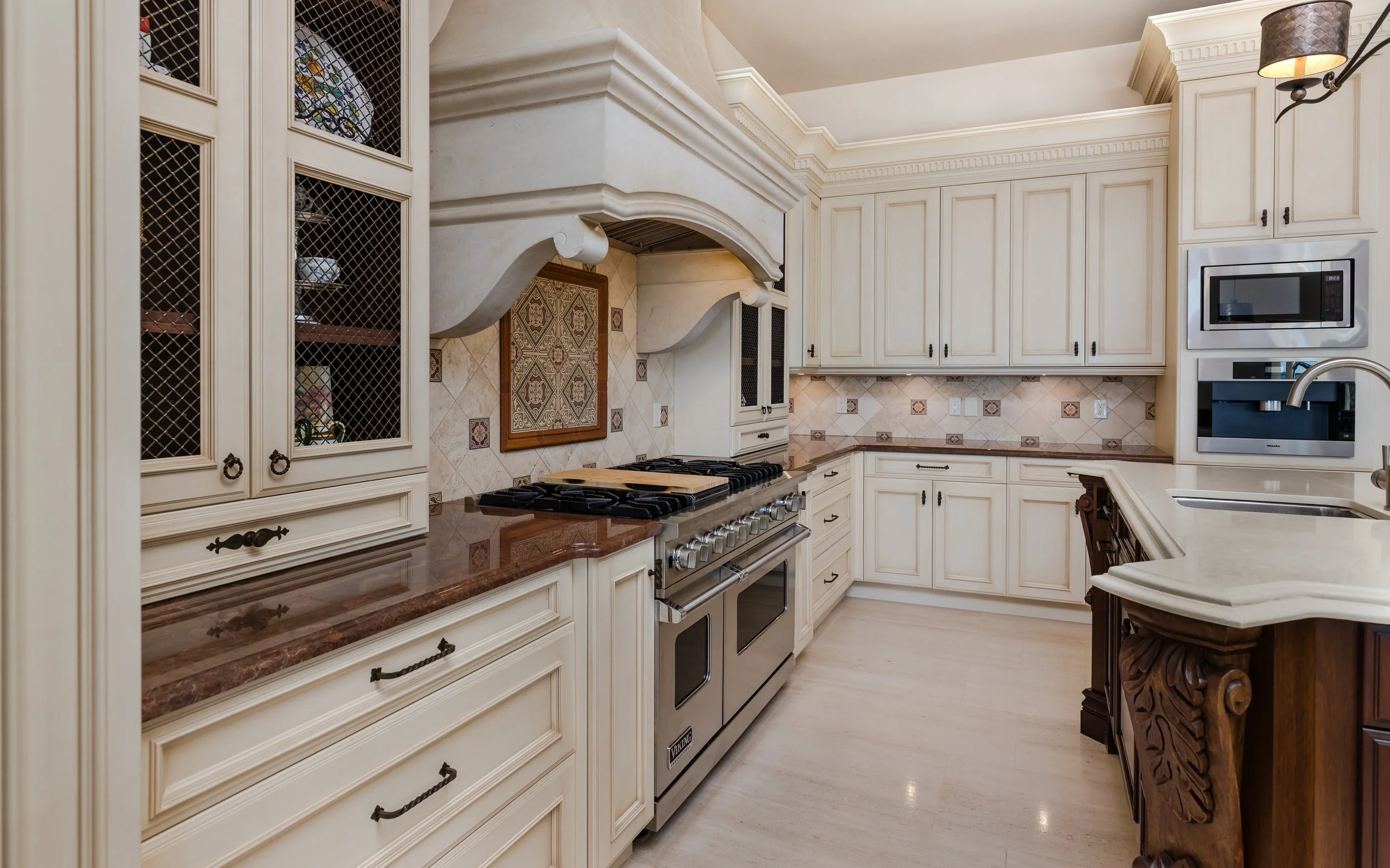 A spacious kitchen with cream-colored cabinets, a patterned tile backsplash, a stainless steel double oven, microwave, coffee maker, and a decorative wooden corner cabinet with carved details.