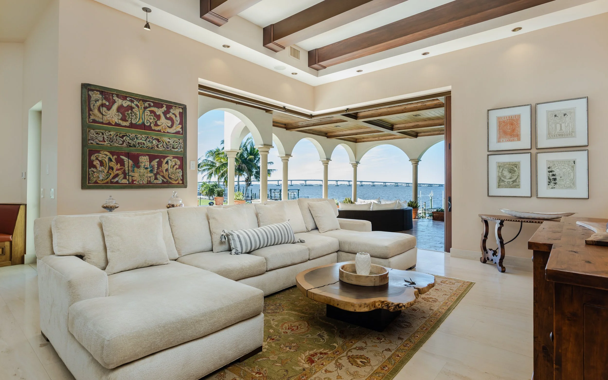 Bright living room with a large beige sectional sofa, wooden coffee table, and view of water and bridge through arches leading to a terrace.