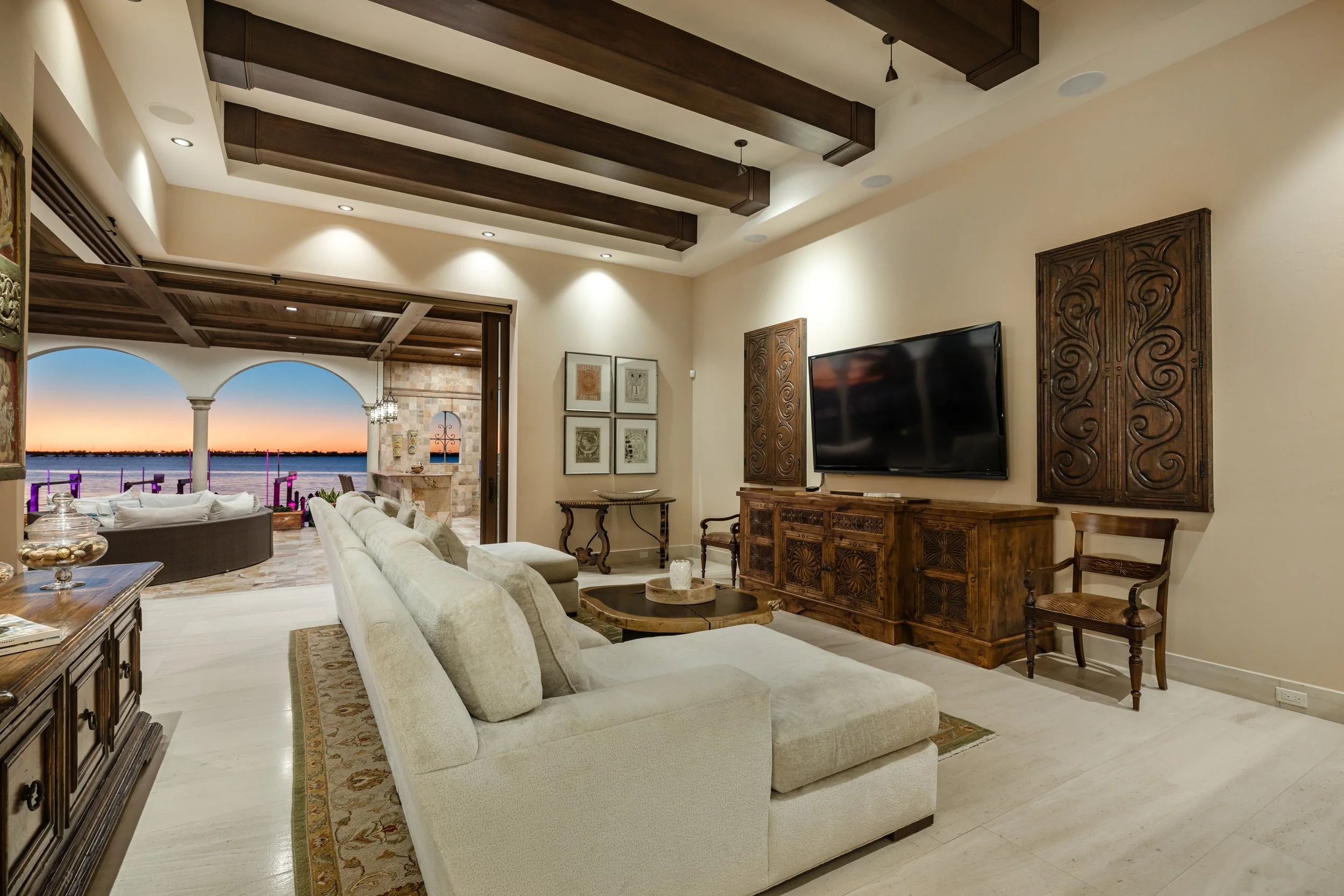 Living room with beige sectional sofa, wooden entertainment center, flat-screen TV, and sliding glass doors opening to a patio overlooking a water sunset view.