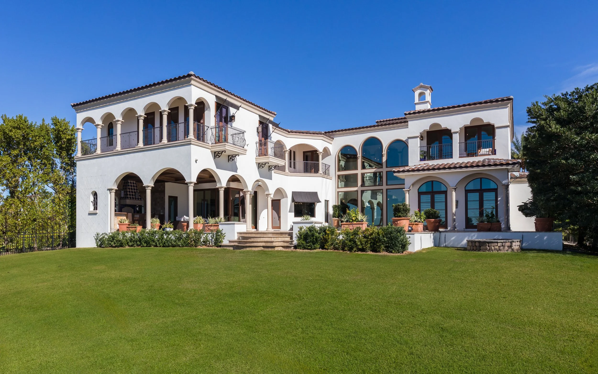 A large Mediterranean-style white villa with multiple balconies, arched windows, and terracotta roof tiles, surrounded by a lush green lawn and trees under a clear blue sky.