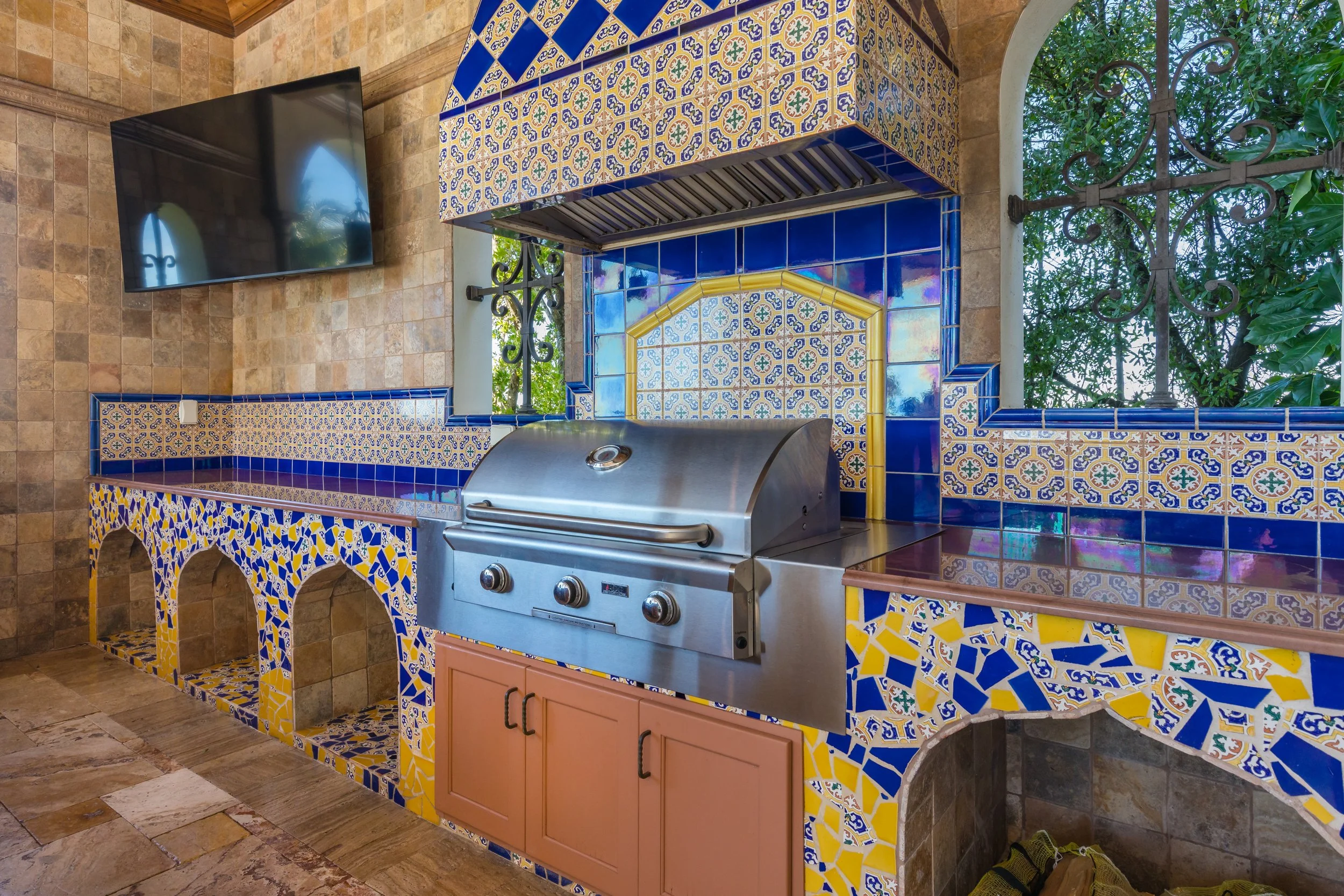 Outdoor kitchen with colorful decorative tiles, stainless steel grill, and flat screen TV on the wall.