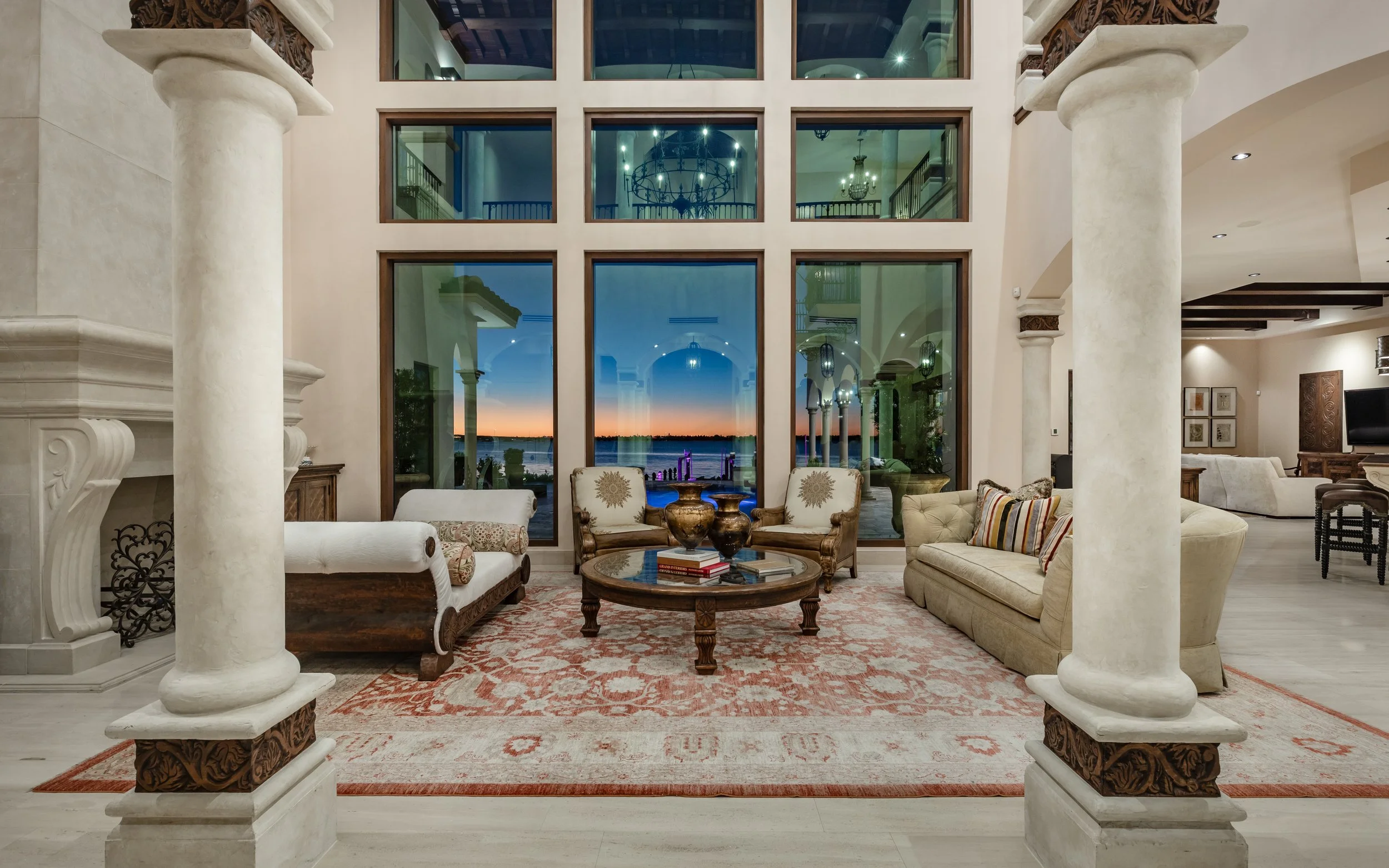 Elegant living room with large windows showing sunset view, classical columns, and vintage furniture.