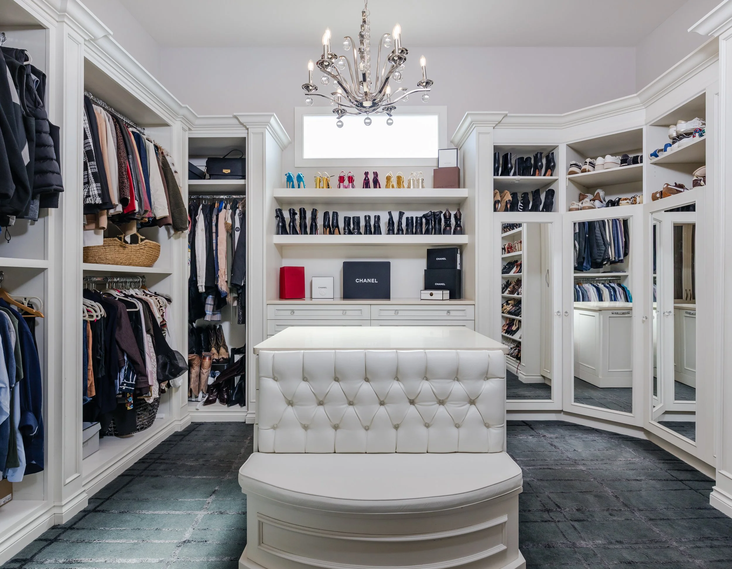 A walk-in closet with white cabinetry, a chandelier, and a white upholstered bench in the center. The closet has shelves filled with clothes, shoes, and accessories, and mirrored doors.