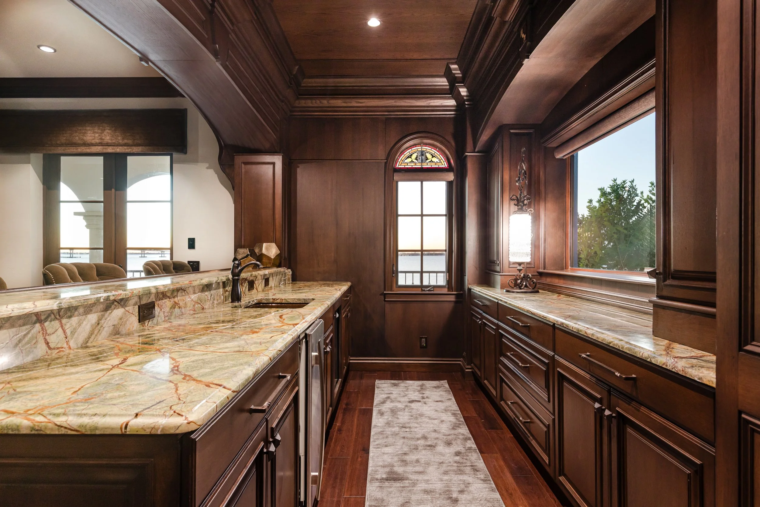 A kitchen with dark wooden cabinets, marble countertops, and a window overlooking greenery and a body of water. The kitchen features a pendant light, hardwood floors, and a small rug.