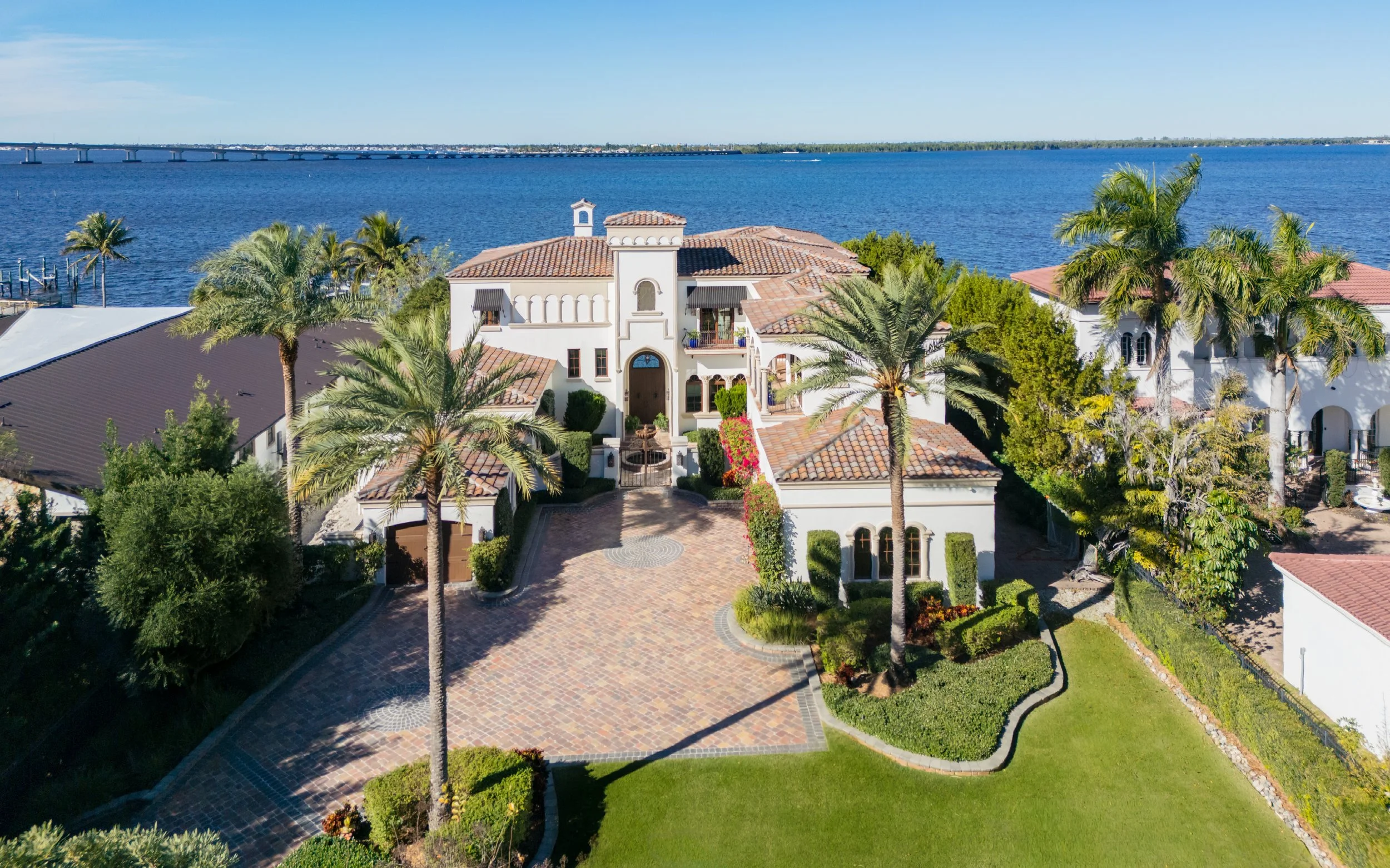 A large Mediterranean-style house with a tile roof, surrounded by palm trees, greenery, and a brick driveway, located near a body of water with a bridge in the background.