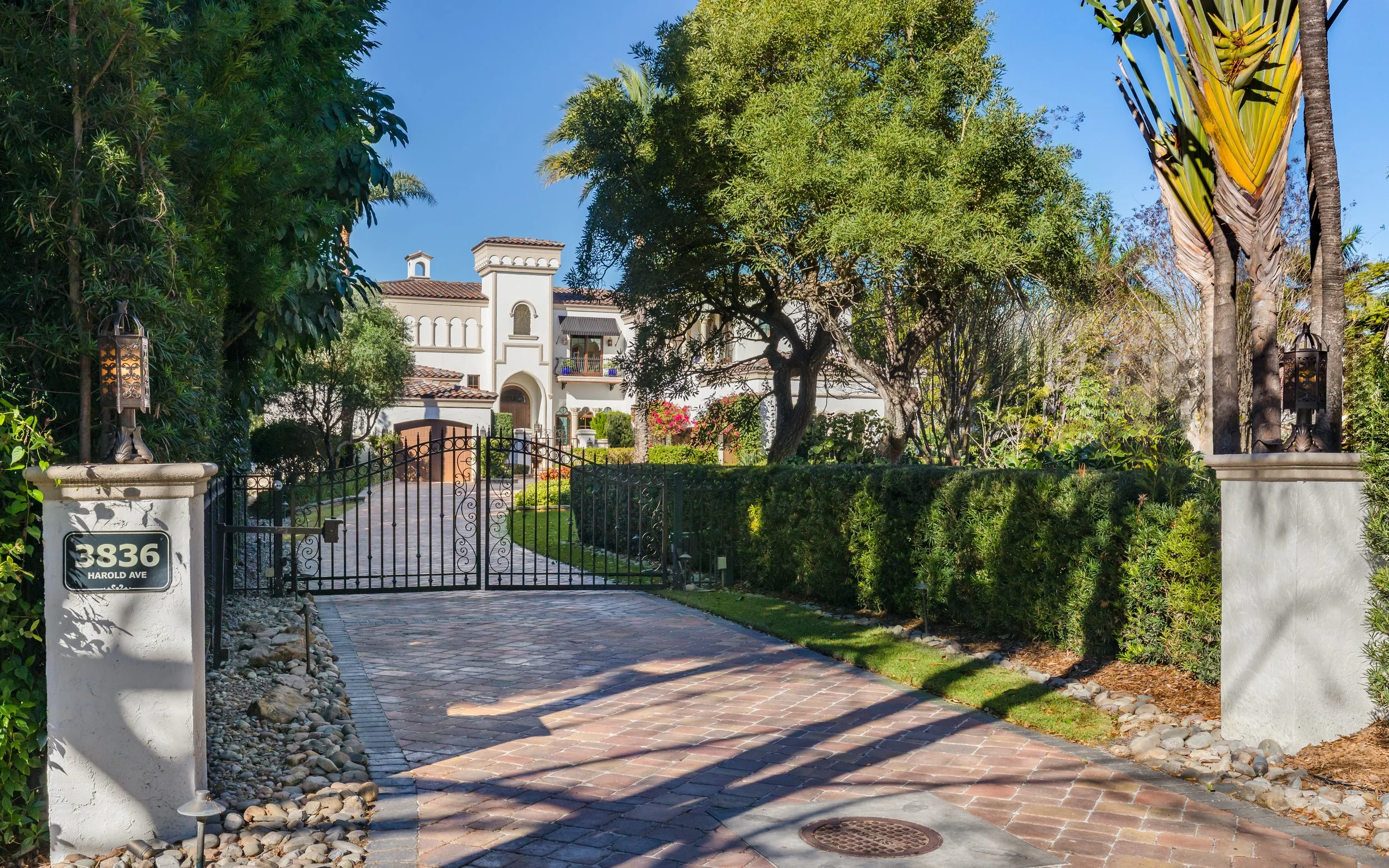 Luxury gated residential property with a brick driveway, black iron gate, lush greenery, trees, and a white multi-story house with balconies.