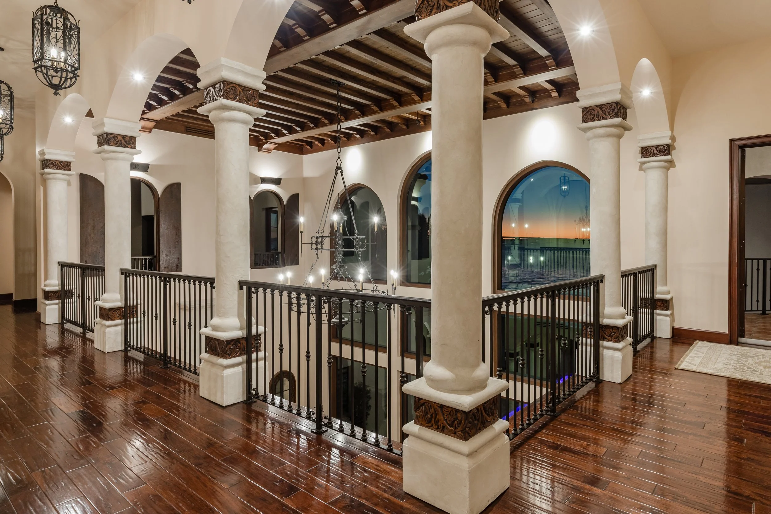 Upscale indoor balcony with wooden floors, white columns with decorative capitals, arched windows showing sunset, wrought iron railing, wooden ceiling, chandelier lighting, and a view down to the lower level.