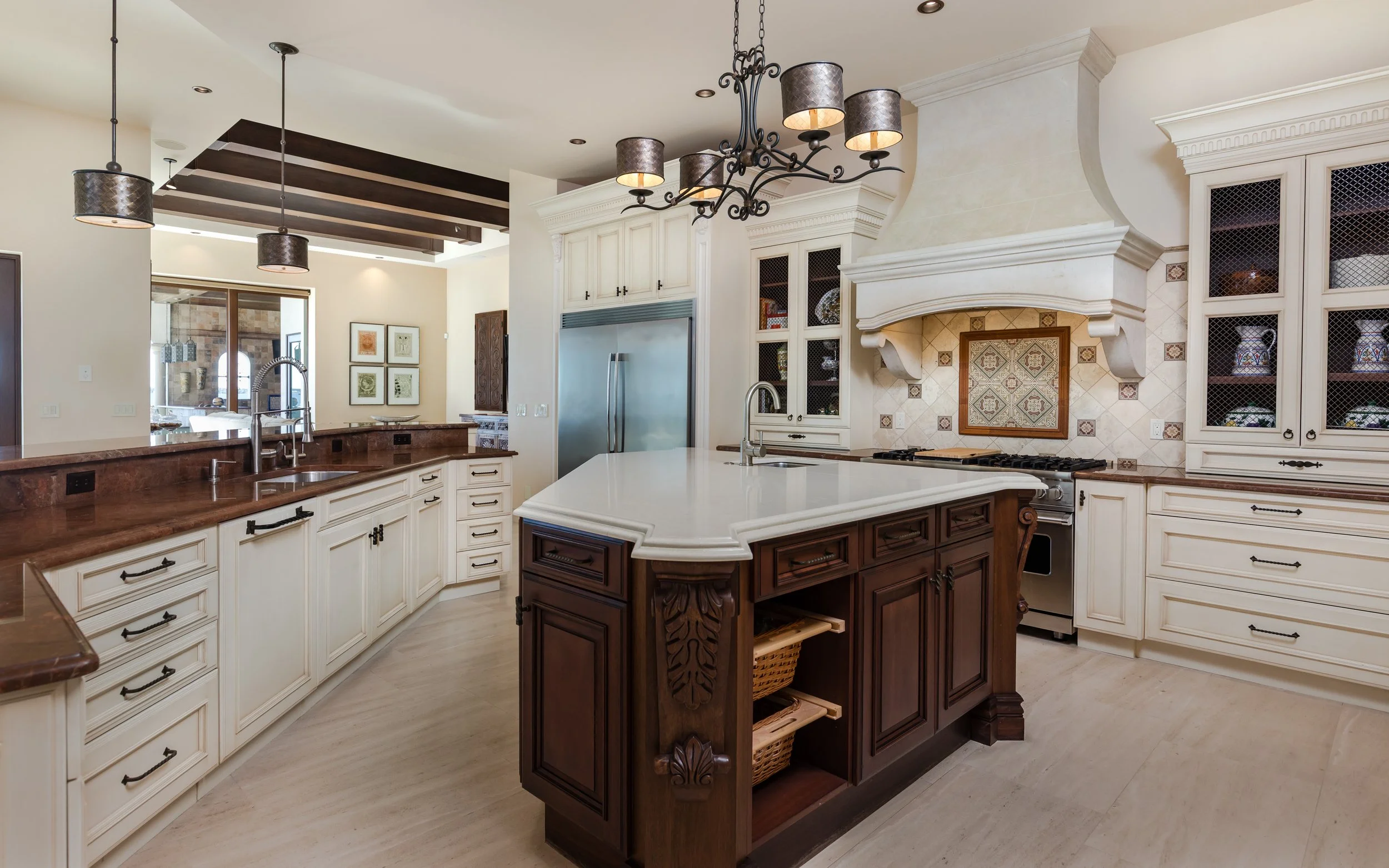 A spacious kitchen with white cabinetry, a large white kitchen island, and dark wood accents. It features multiple hanging pendant lights and a decorative chandelier. There is a stainless steel refrigerator and a traditional stove with a detailed ran