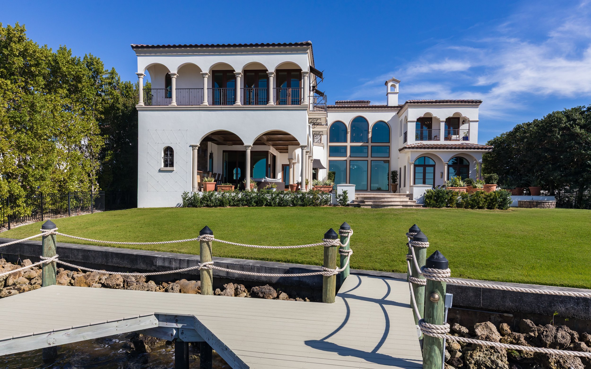 Large white Mediterranean-style house with multiple balconies, large arched windows, and a well-maintained lawn, located near water with a dock and rope railing.