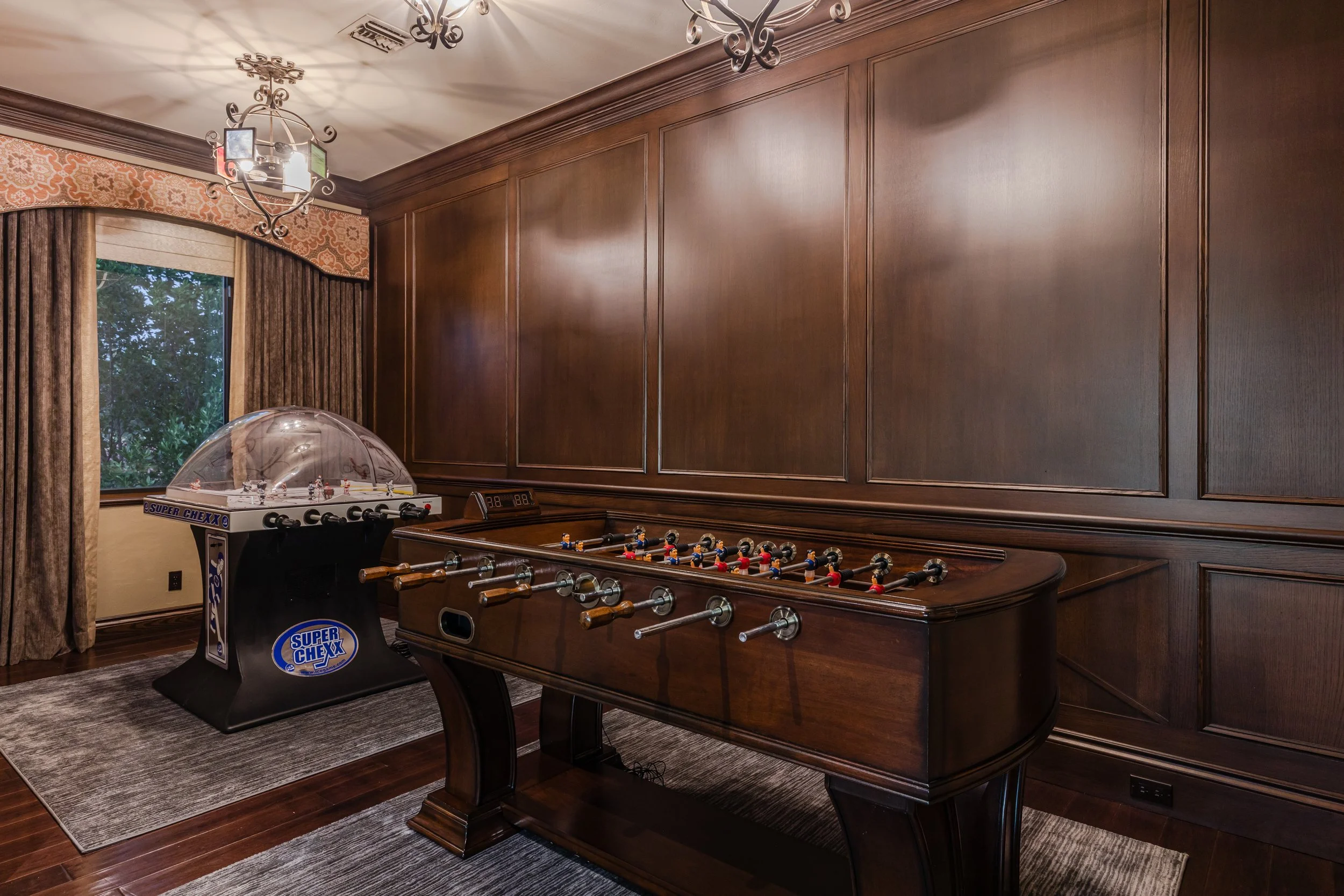 A room with dark wooden wall paneling and flooring, containing a foosball table and an air hockey table, with a window showing trees outside, curtains, and two decorative chandeliers.