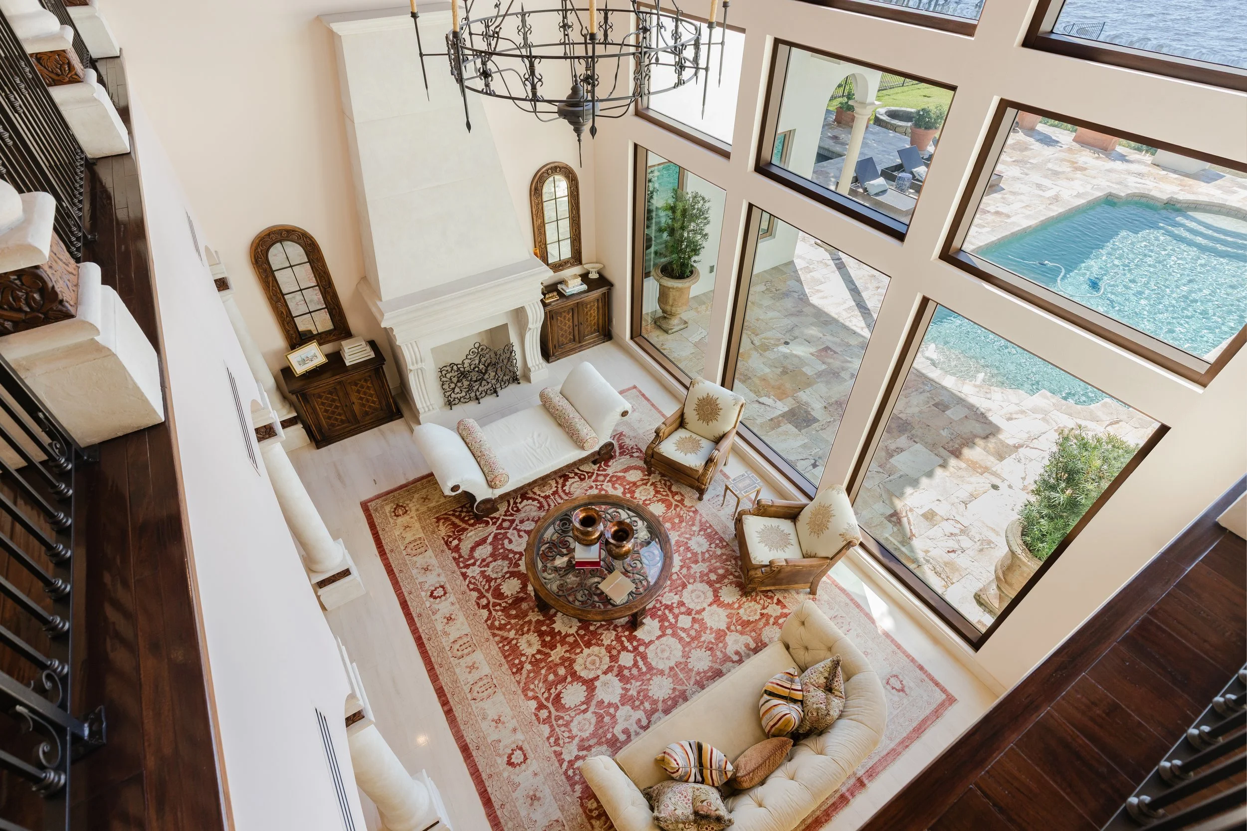 A spacious living room with large floor-to-ceiling windows overlooking a swimming pool and patio area. The room features a cream-colored sofa, two matching armchairs with patterned cushions, a round coffee table with decorative items, and a red patte