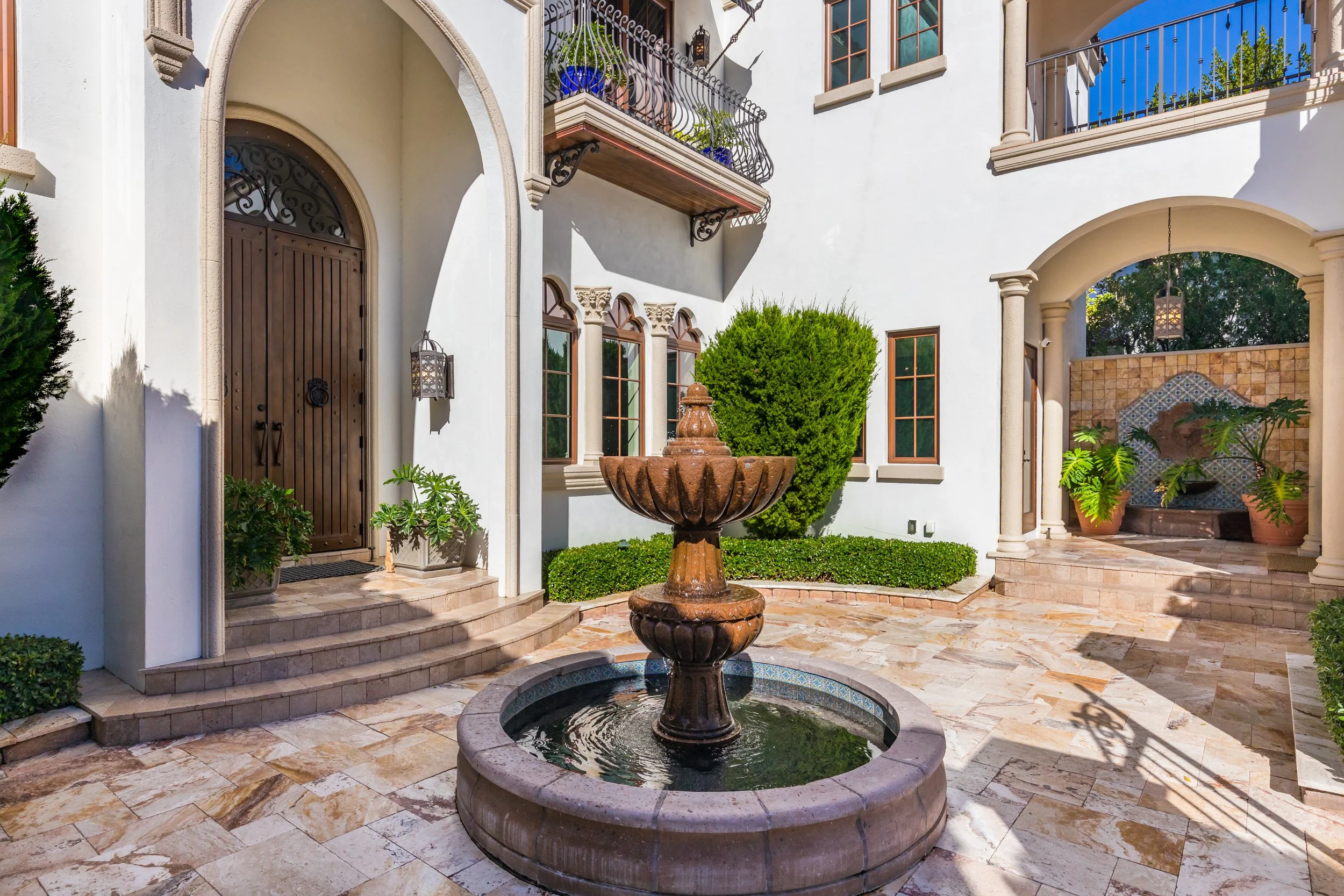 Courtyard of a luxurious house with a stone fountain, tiled floor, bushes, and balconies with iron railings.