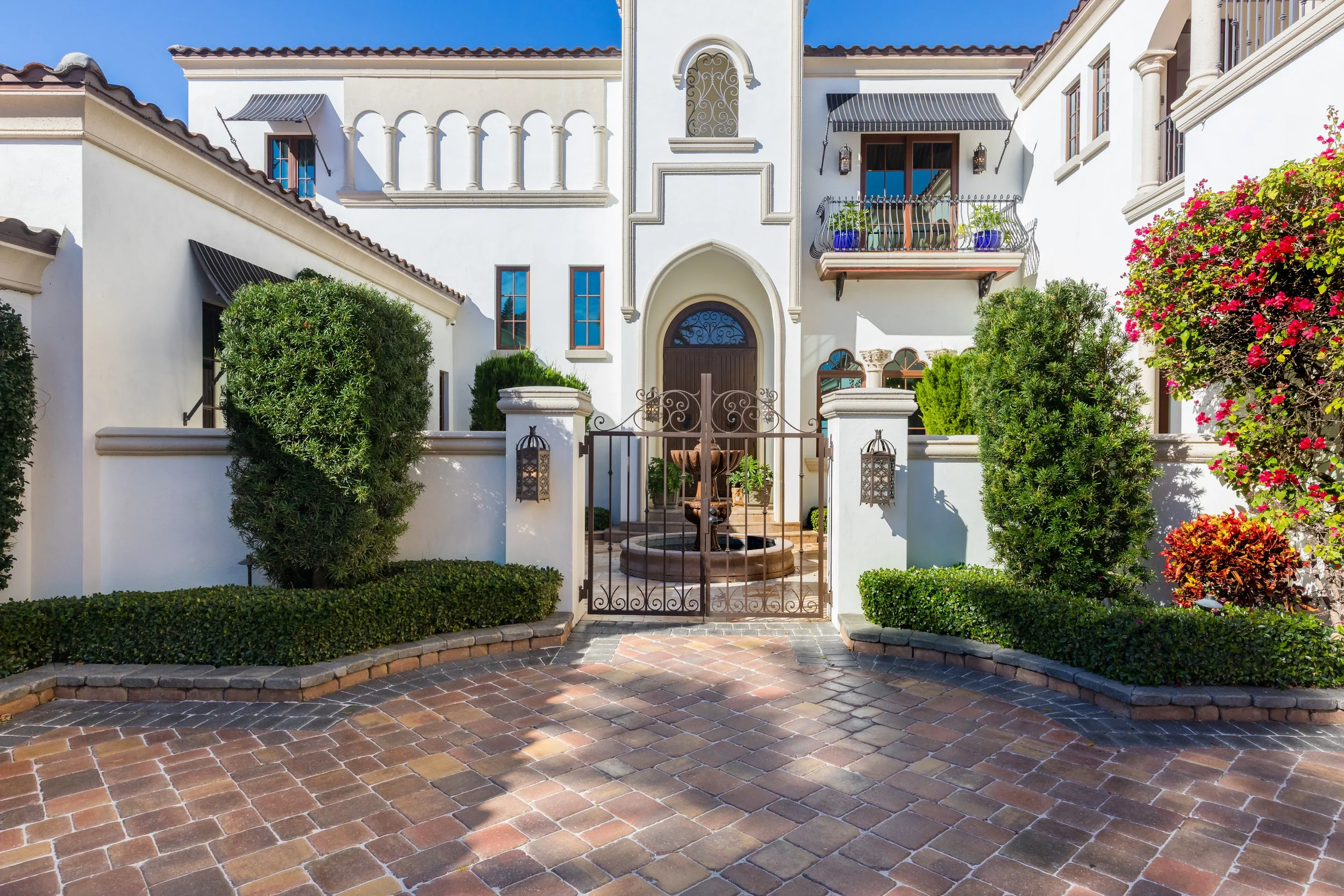 Front view of a white Mediterranean-style house with a wrought iron gate, brick pathway, lush green bushes, and blooming flowers under a bright blue sky.