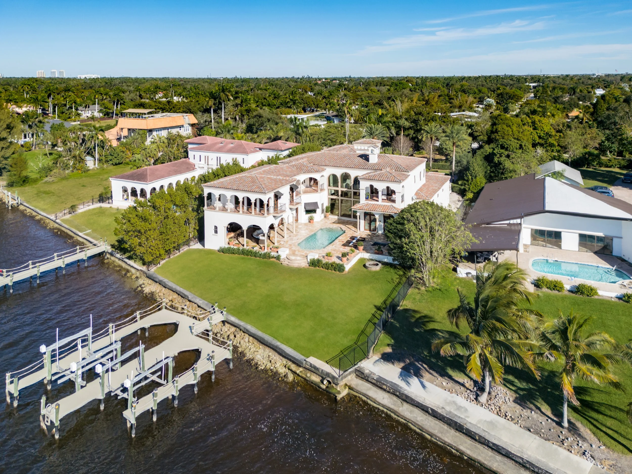 A large white waterfront house with multiple patios, a swimming pool, and a dock on the water, surrounded by greenery and neighboring homes.
