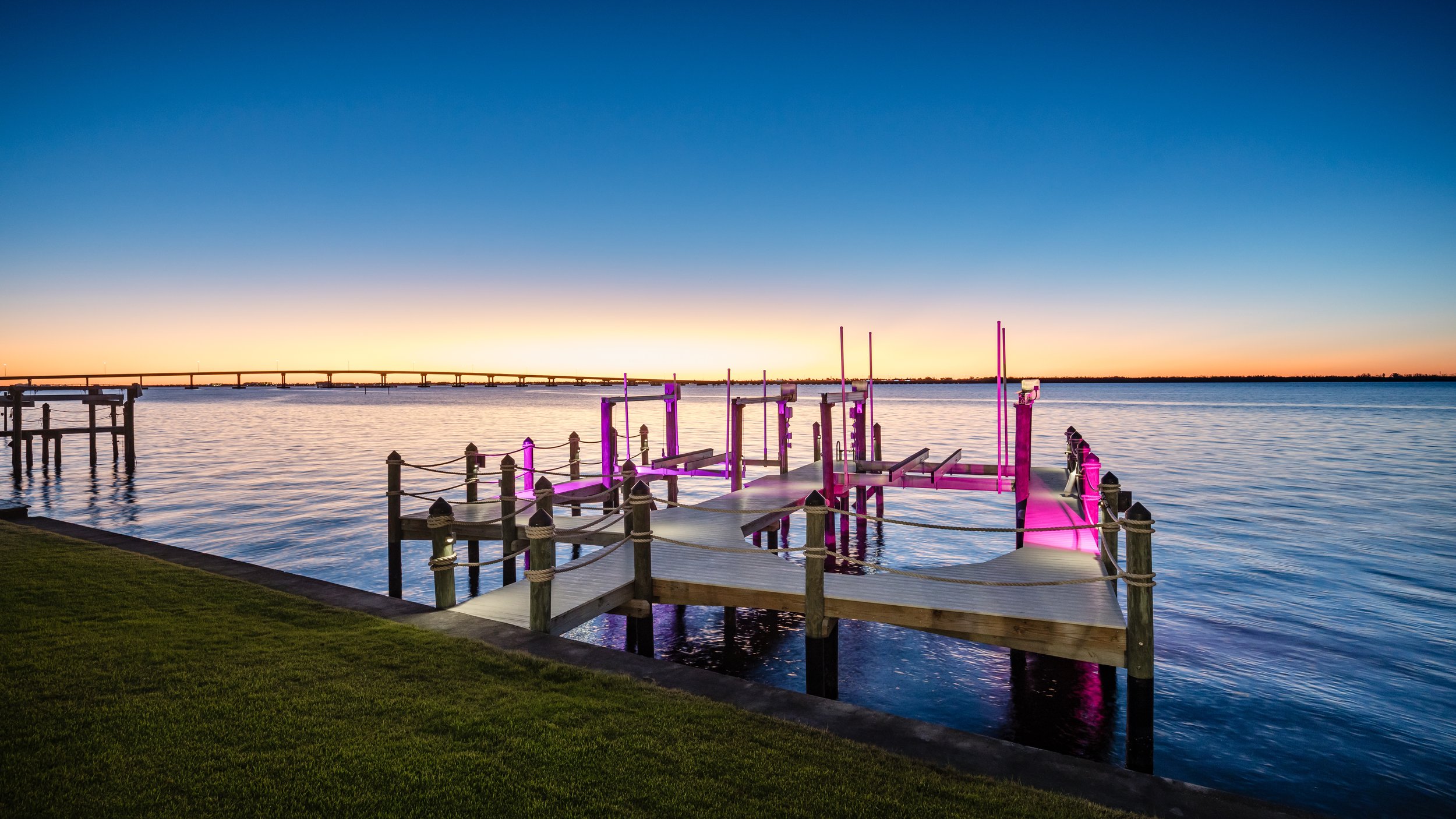 A wooden dock extending into a calm lake with pink LED lights along the edges, against a sunset sky with a distant bridge.