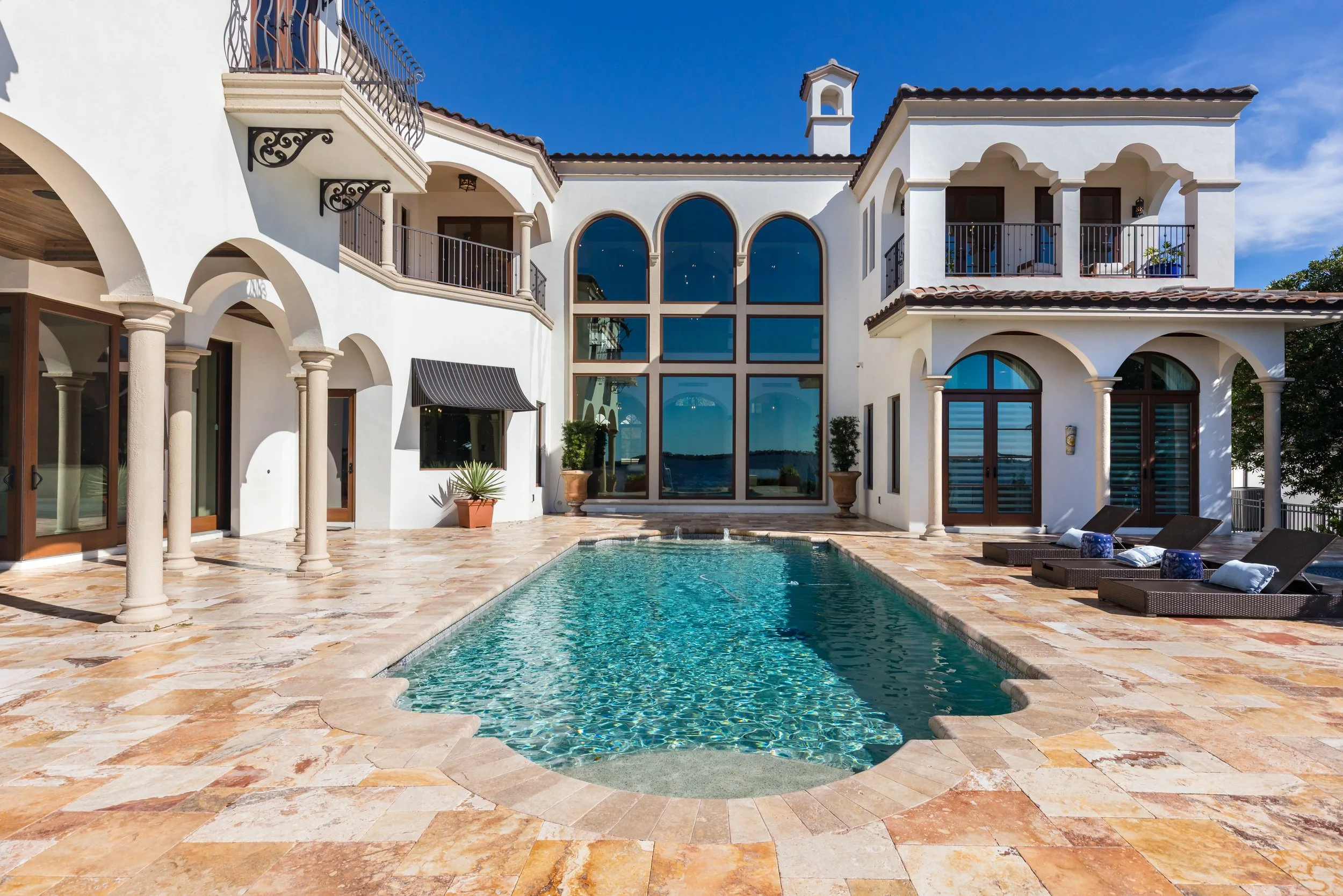 Luxury white villa with arched windows, balconies, and a swimming pool in front on a sunny day.