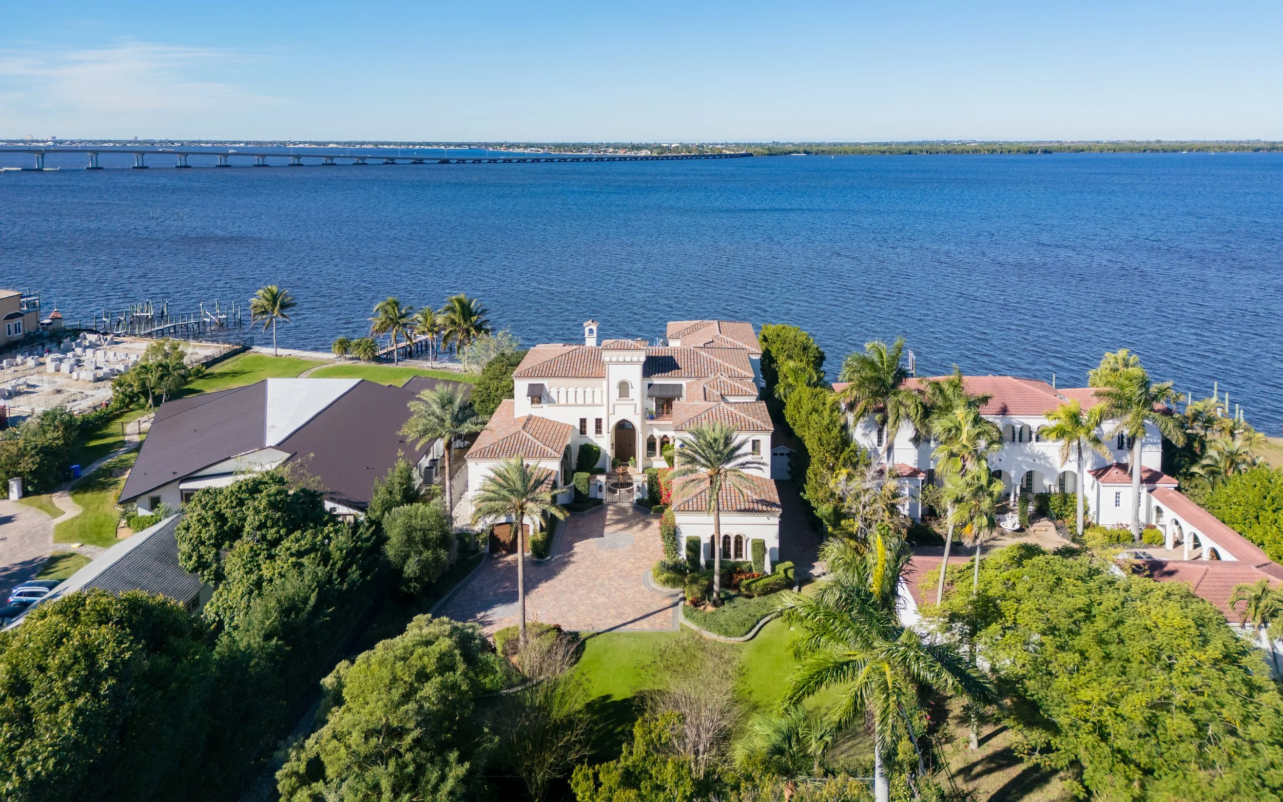 Aerial view of a large luxurious waterfront estate with palm trees, a drive-in courtyard, and neighboring houses, overlooking a wide river or lake.