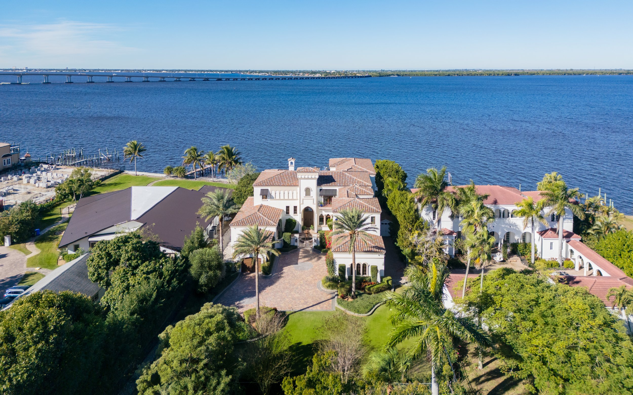 Aerial view of luxury waterfront homes surrounded by lush greenery, overlooking a wide river with a bridge in the distance, under a clear blue sky.