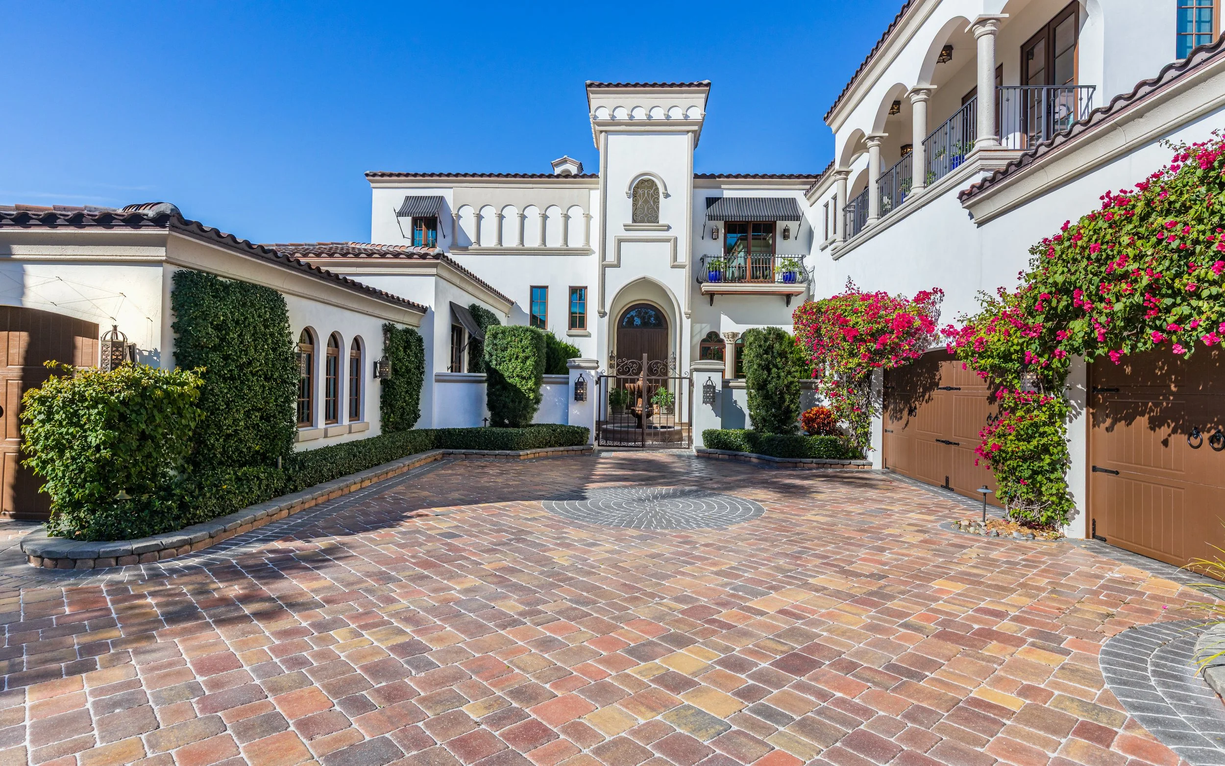 Large Mediterranean-style white house with brown tiled roof, courtyard with brick pavers, and lush greenery including flowering bushes, with a black gate and blue sky in background.