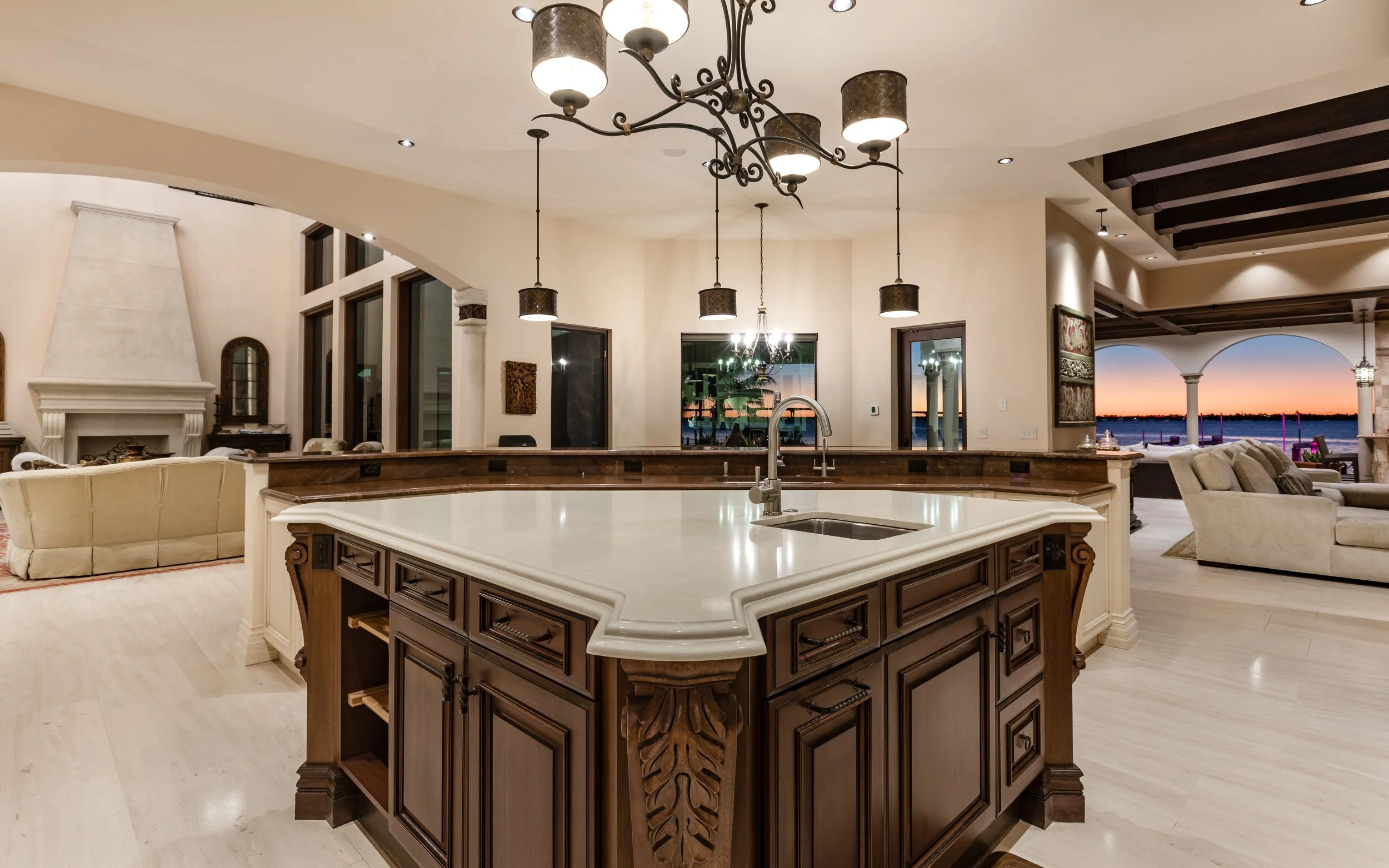 A large open-concept kitchen and living room area with a central kitchen island featuring a white countertop, dark wooden cabinetry, and pendant lighting fixtures overhead. The space is illuminated with warm, ambient lighting, and there are views of 