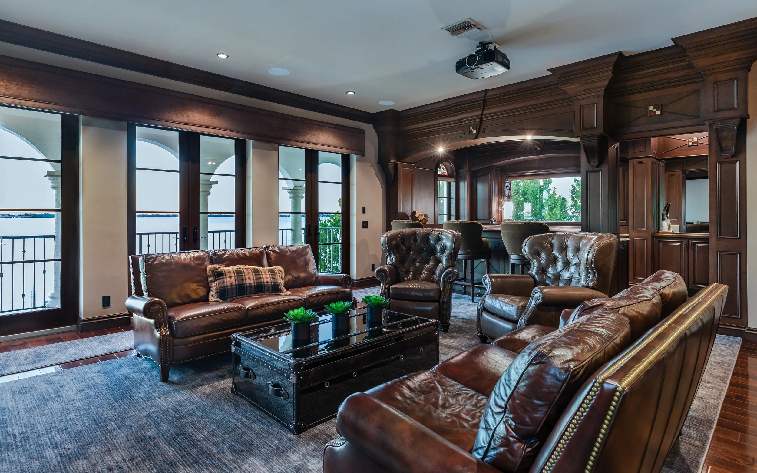 Luxurious living room with leather sofas, armchairs, large windows, and a bar area with barstools, dark wood paneling, and a view of trees outside.