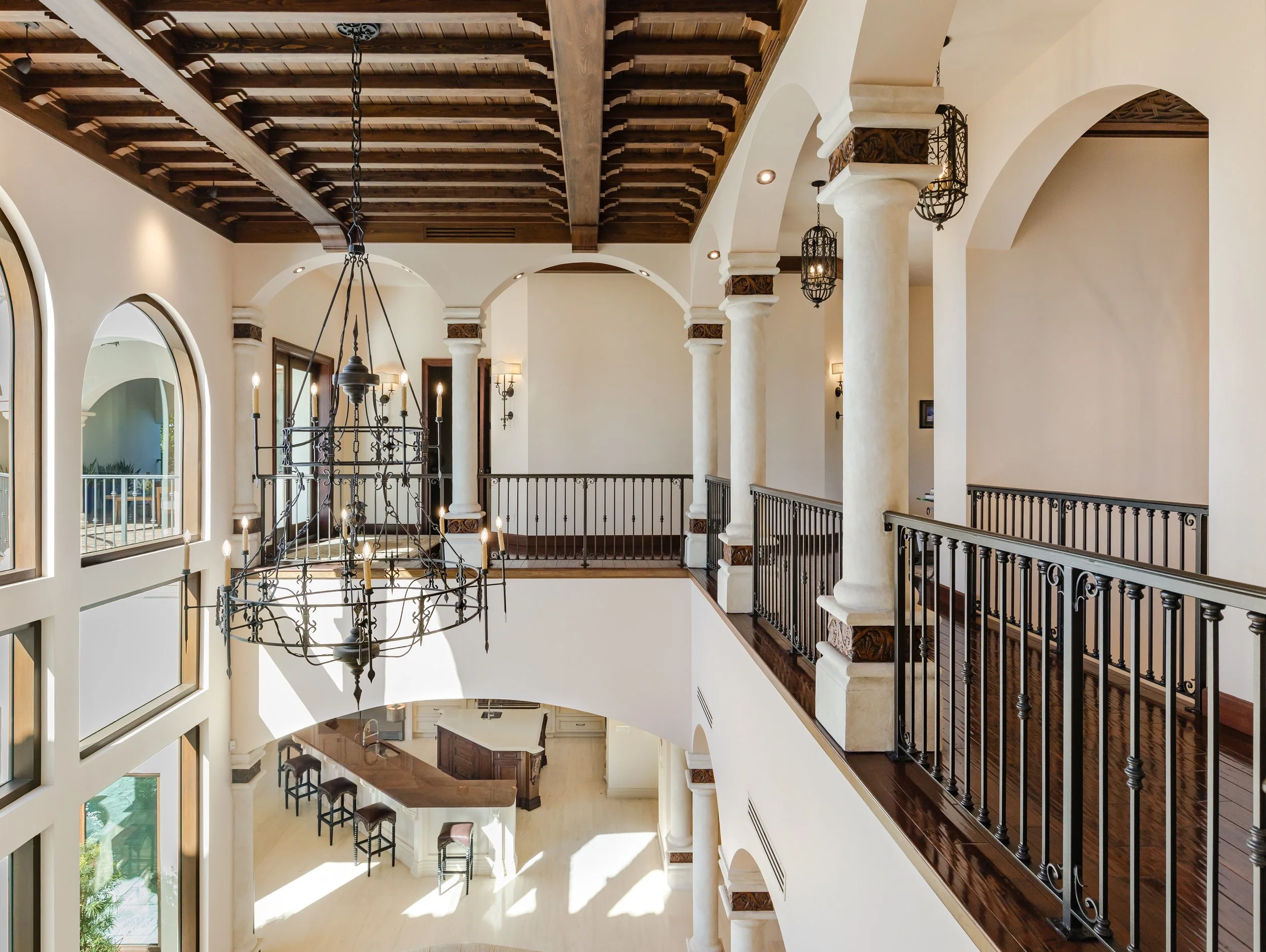 Interior view of a spacious, upscale house with a high ceiling, large arched windows, a wrought iron chandelier, and a mezzanine with iron railings, overlooking the living area below.