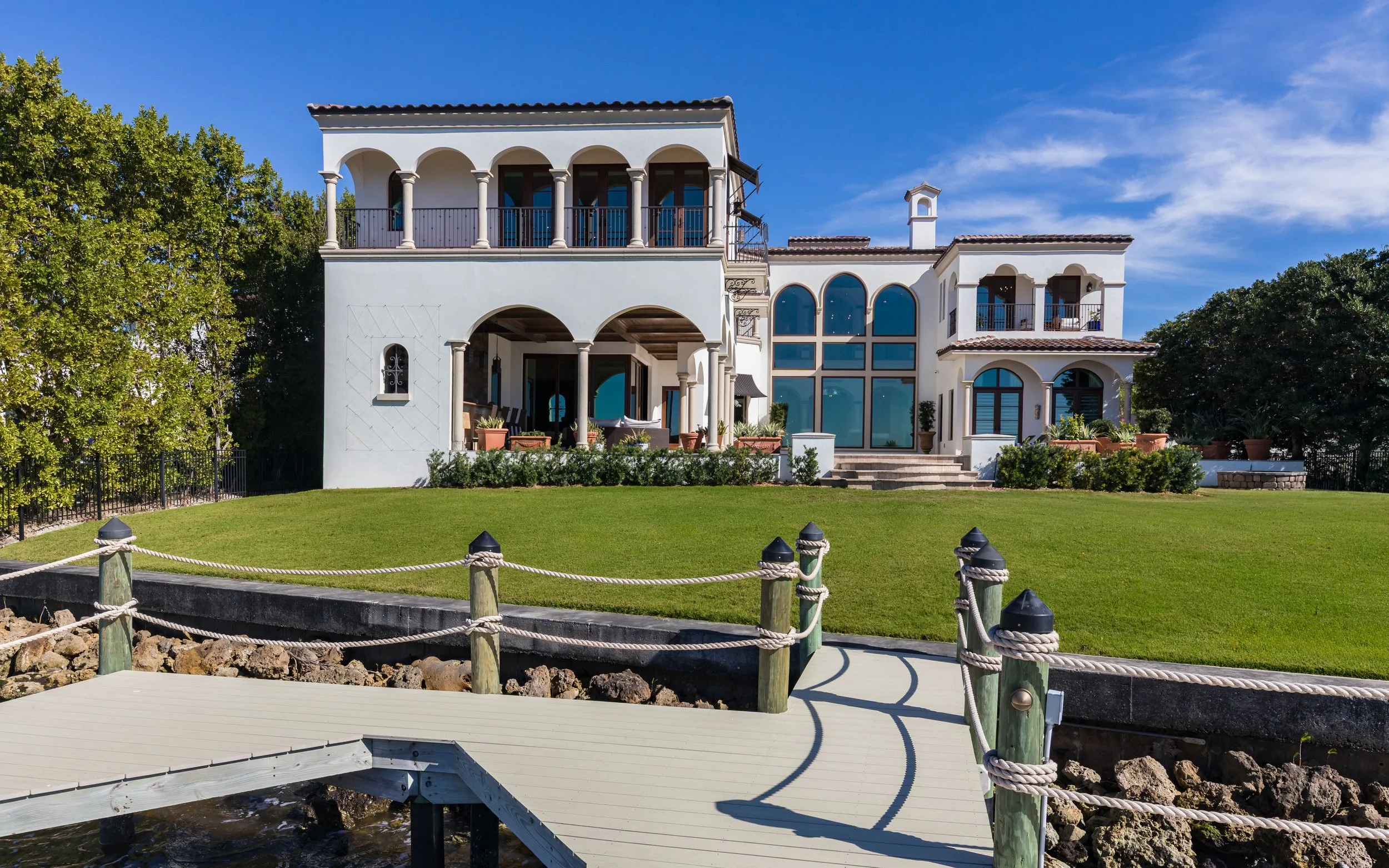 A large, white Mediterranean-style house with multiple balconies and arched windows, overlooking a grassy yard with a dock near water.