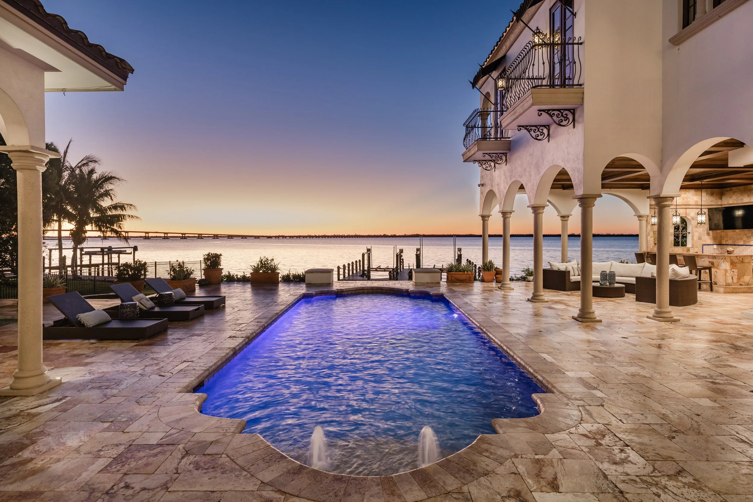 Luxury backyard with a swimming pool overlooking a lake at sunset, surrounded by lounge chairs, potted plants, and a covered patio with outdoor seating and a bar area.