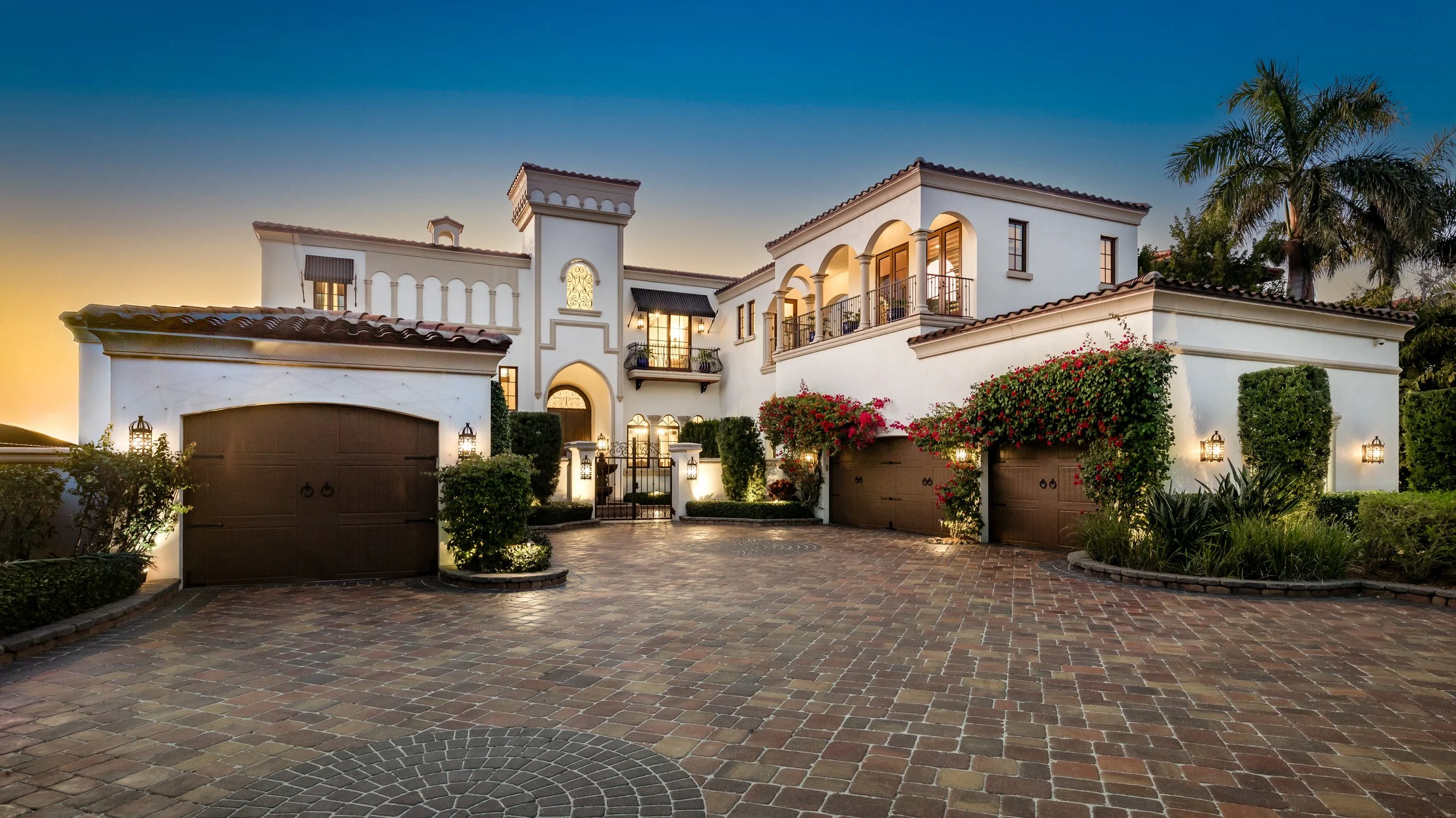 A large, elegant white house with multiple stories, arched windows, and balconies. It features a gated entrance, surrounding greenery, and a paved driveway with street lamps and flowering bushes.