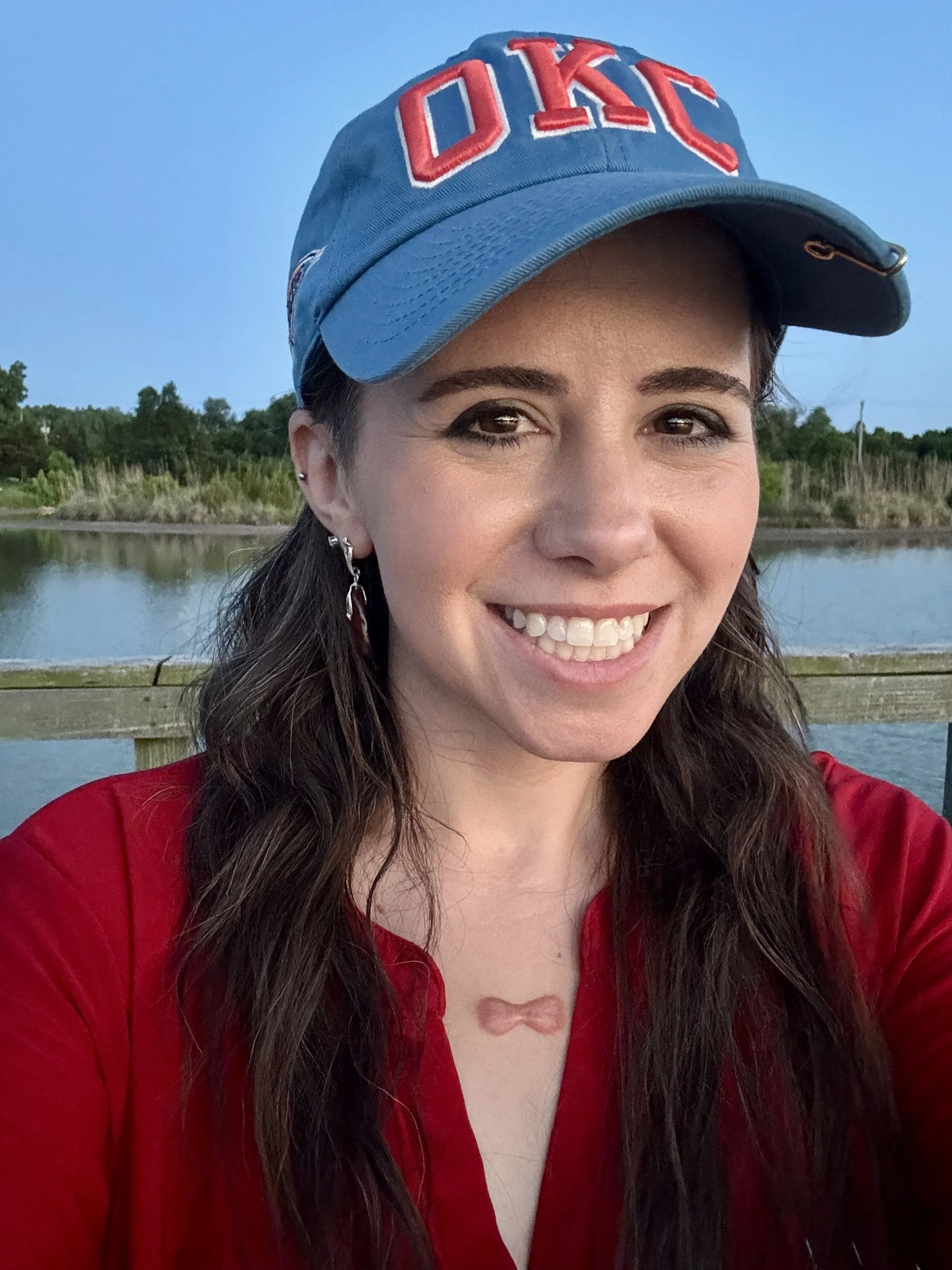 A smiling woman wearing a red shirt and a blue cap with red lettering, standing outdoors by a body of water with trees in the background.