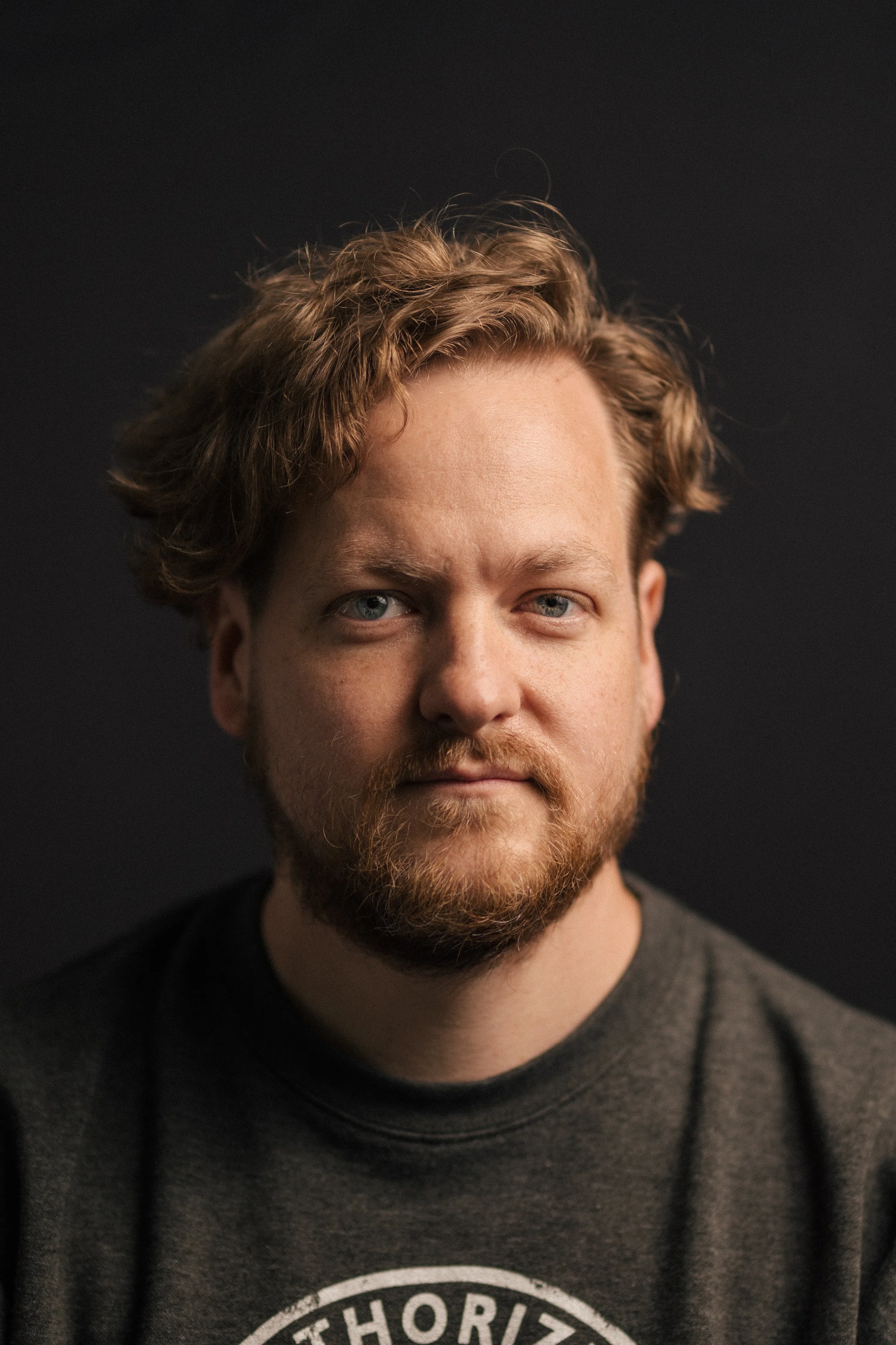 Close-up of a man with curly blonde hair and a beard, wearing a dark gray sweatshirt, against a black background.