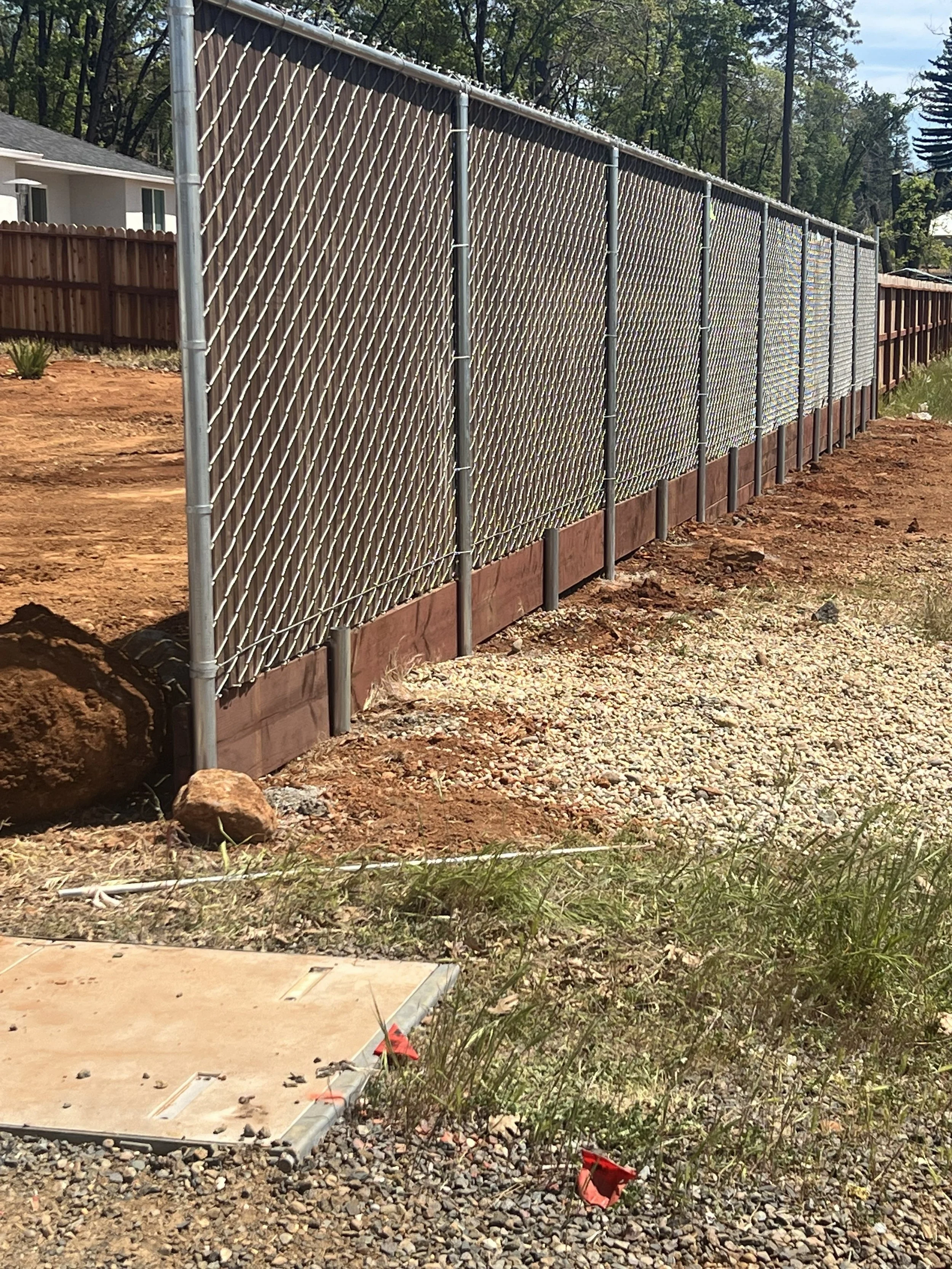 A long metal chain-link fence with wooden panels at the bottom, set on a dirt and gravel ground.