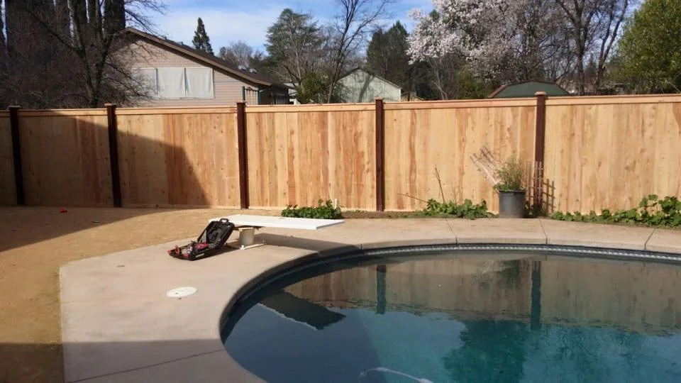 Residential backyard with a concrete pool, wooden privacy fence, tools and equipment on the pool deck, and some plants and a potted shrub near the fence. The sky is partly cloudy.