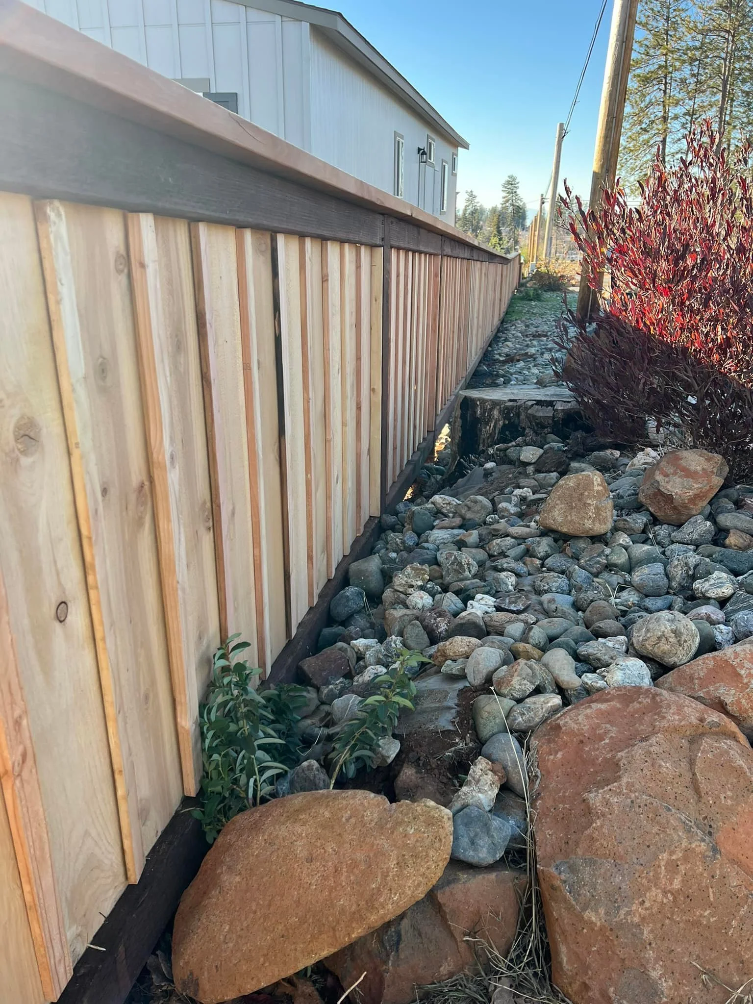 A wooden privacy fence runs alongside a house with a white exterior, set in a rocky landscape with large stones and some green plants. A few red-leaved bushes and trees can be seen in the background, along with utility poles and lines under a clear blue sky.