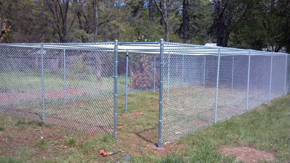 A long chain-link fence with a gated entrance, set on a grassy area with trees in the background.