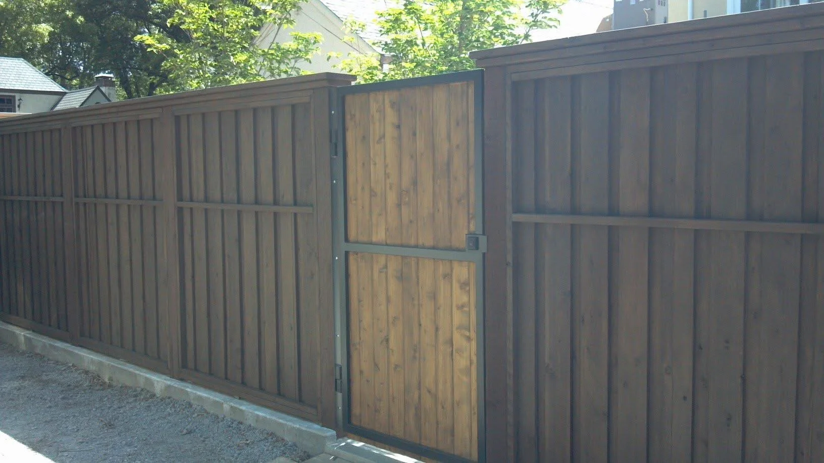 A wooden privacy fence with a small gate in the center, set against a background of trees and houses, with sunlight filtering through the leaves.