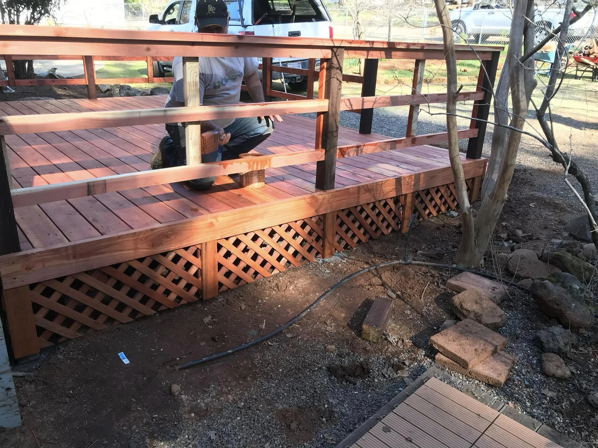 A person kneeling on a wooden deck under construction, with railings being assembled and trees nearby.