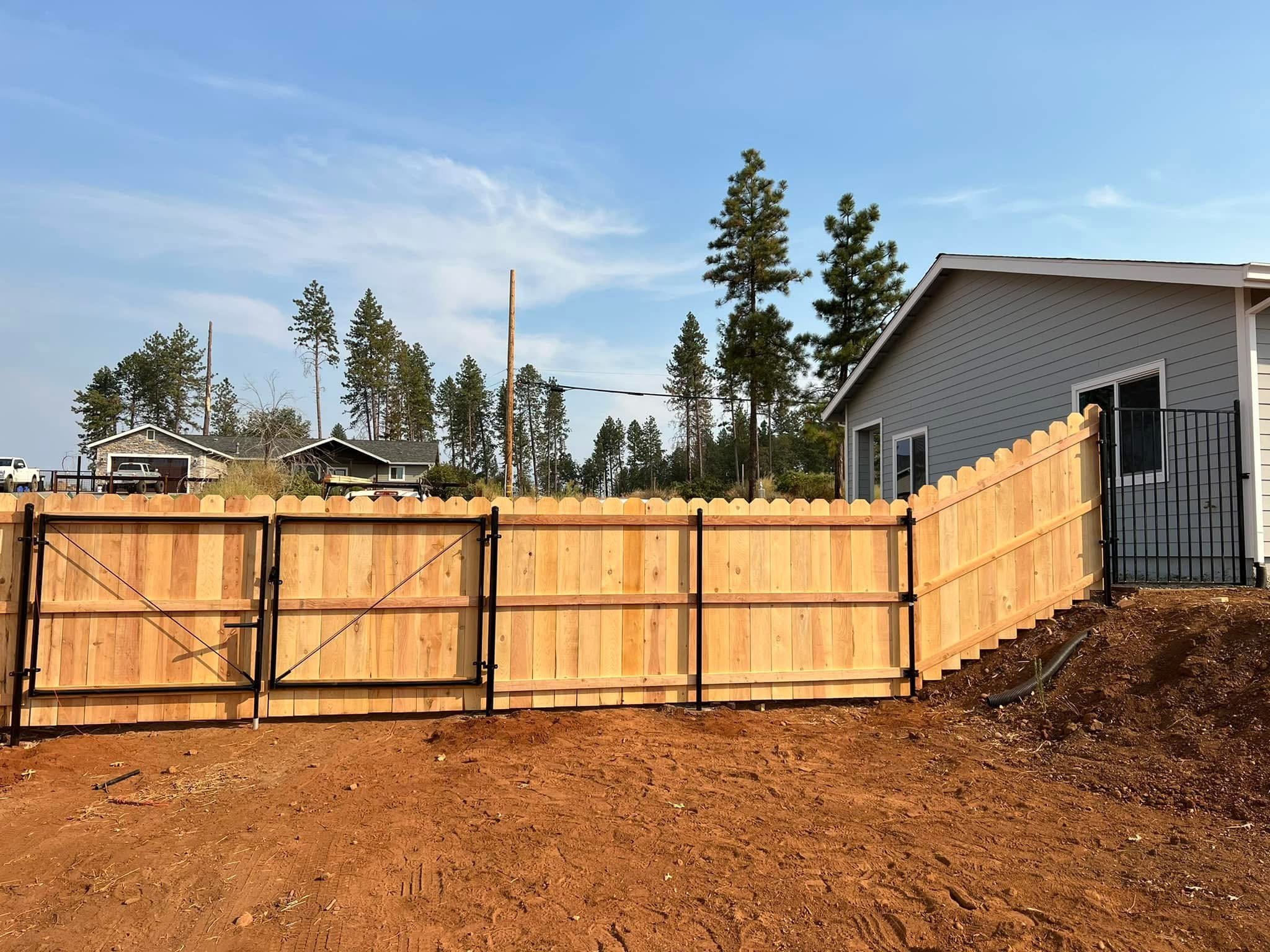 New wooden fence partially installed in front of a house with gray siding, with trees and neighboring houses in the background under a clear sky.