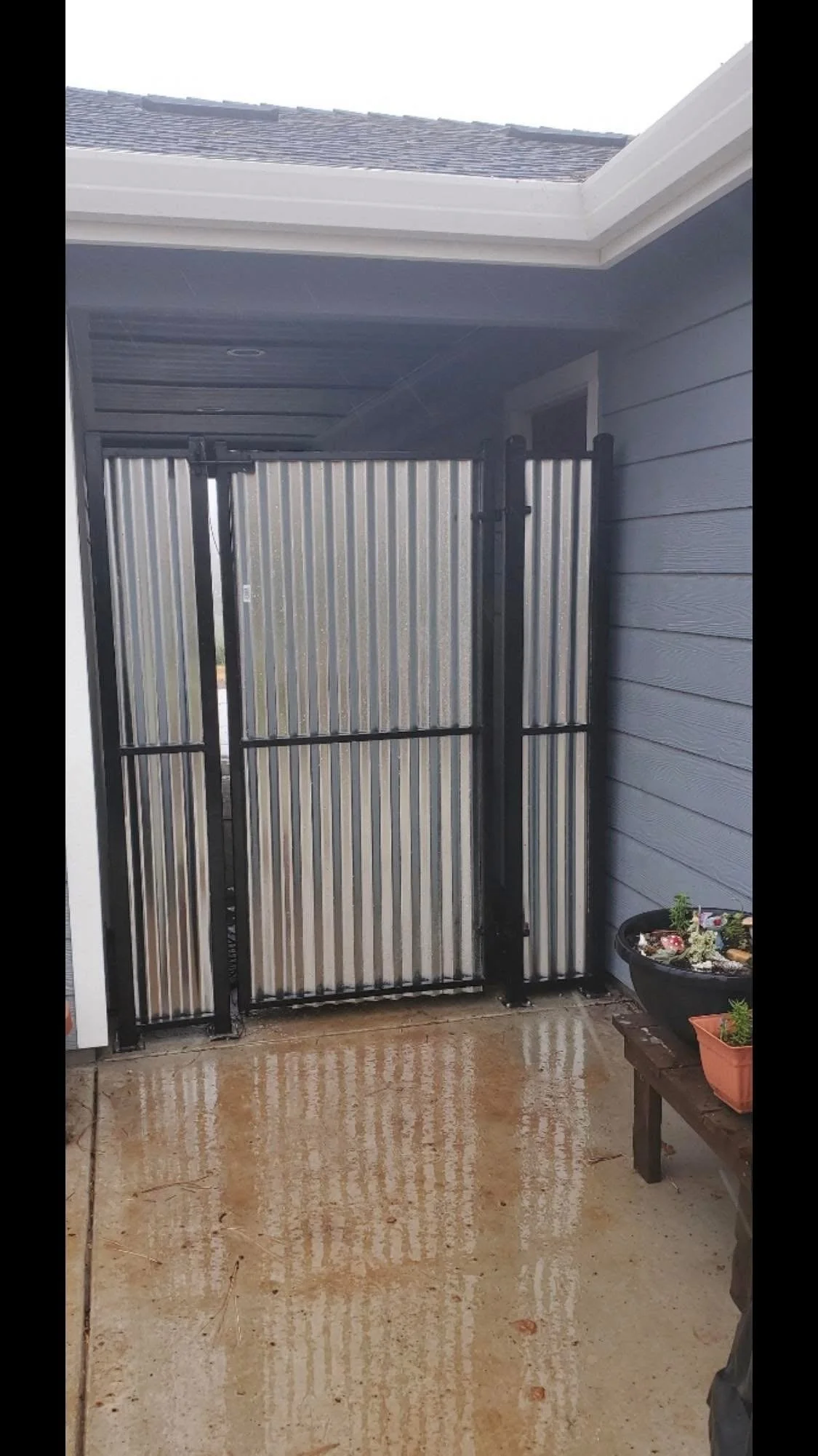 Rainy day view of a metal gate in a porch with a concrete floor, a gray house wall on the right, and a small wooden table with potted plants.