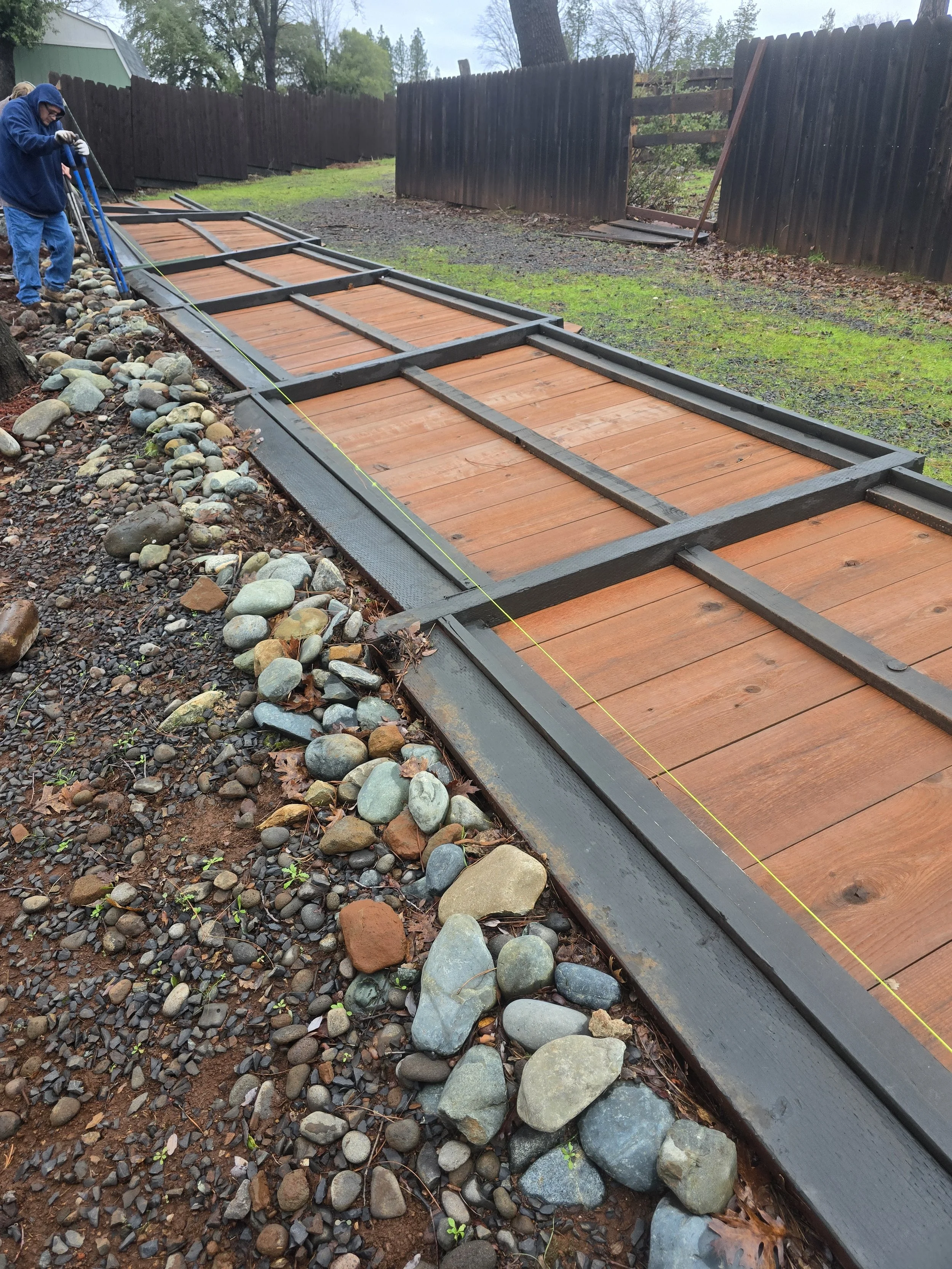 A person installing a wooden pathway with black metal framing in a backyard, with a string line for alignment, rocky ground on the side, and a wooden fence in the background.