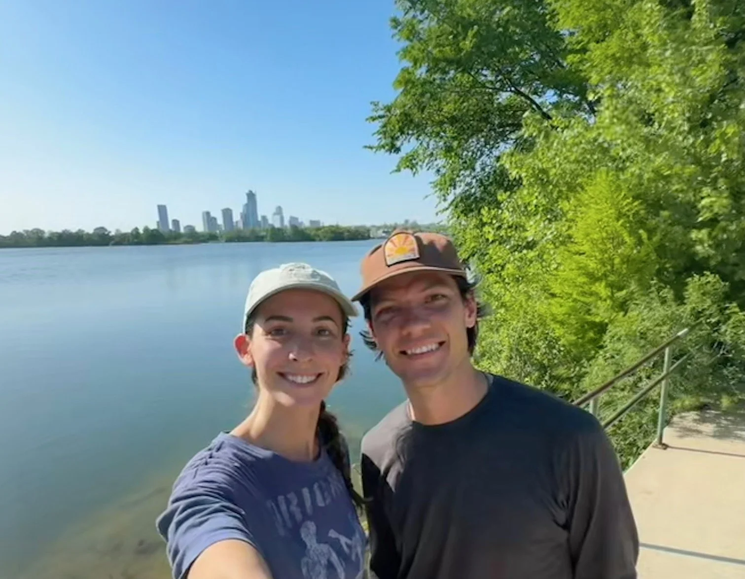 A man and woman taking a selfie outdoors near a body of water, with city skyline in the distance and green trees on the right.