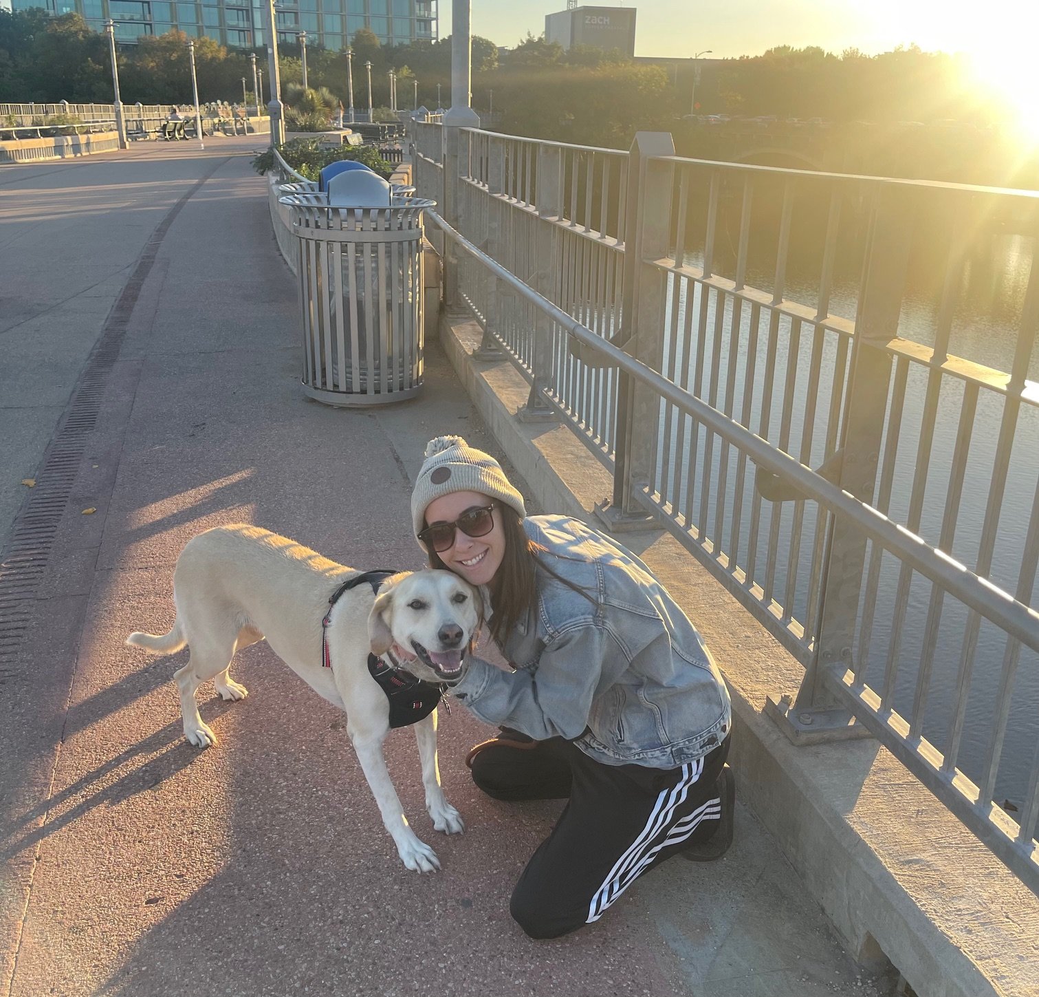 A woman kneeling and smiling with her dog on a paved riverfront walkway during sunset. The woman is wearing sunglasses, a beanie hat, a denim jacket, and black pants with white stripes. The dog is tan and white and is wearing a harness.