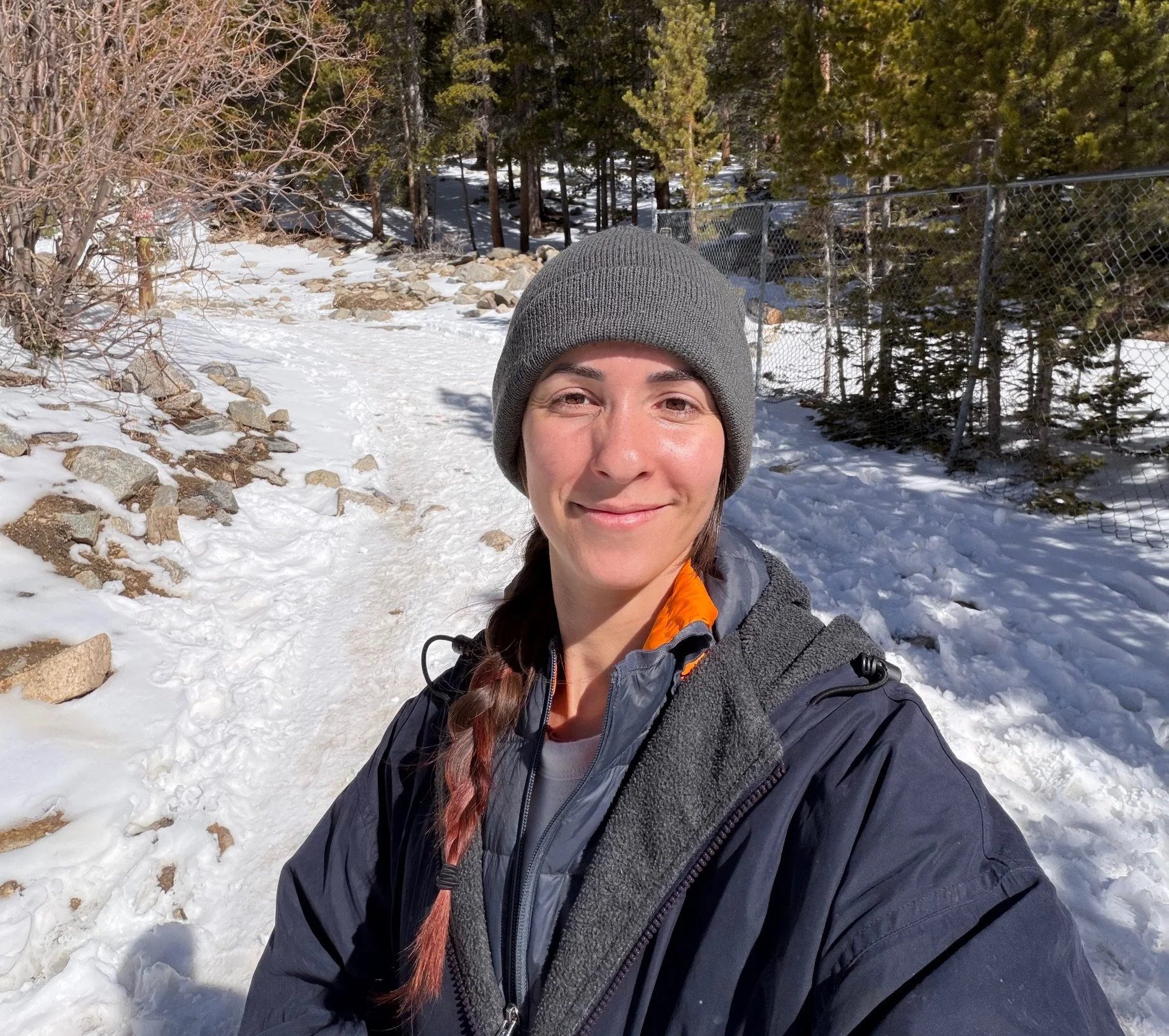 A woman outdoors on a snowy trail, wearing a gray beanie, a black jacket, and a gray fleece, smiling at the camera with a forest background.