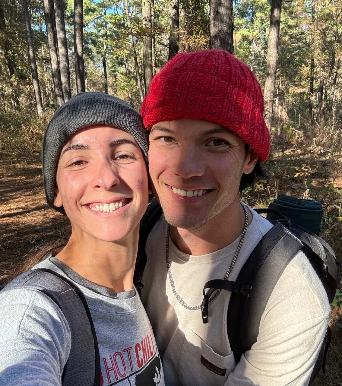 A smiling young woman and man taking a selfie in a forest with tall trees, both wearing beanies and casual clothes, carrying backpacks.