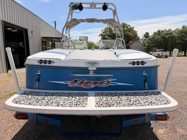 A blue and white Triton boat on a trailer, parked outdoors near a building, with trees and other vehicles in the background.